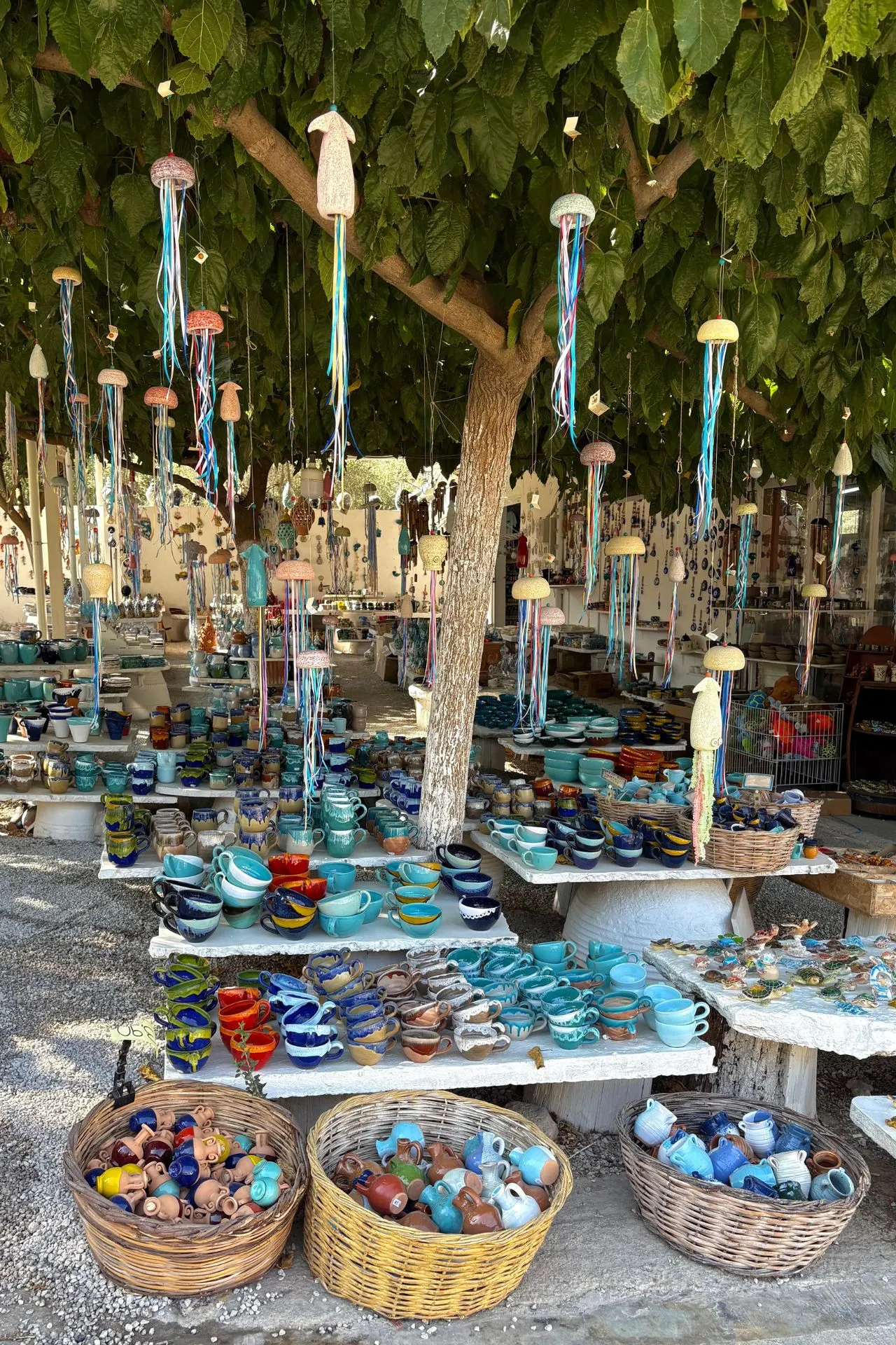 Kleurrijke keramiekmarkt onder een boom vol hangdecoraties, met schalen, mokken en kommen in turquoise, blauw en aardetinten.