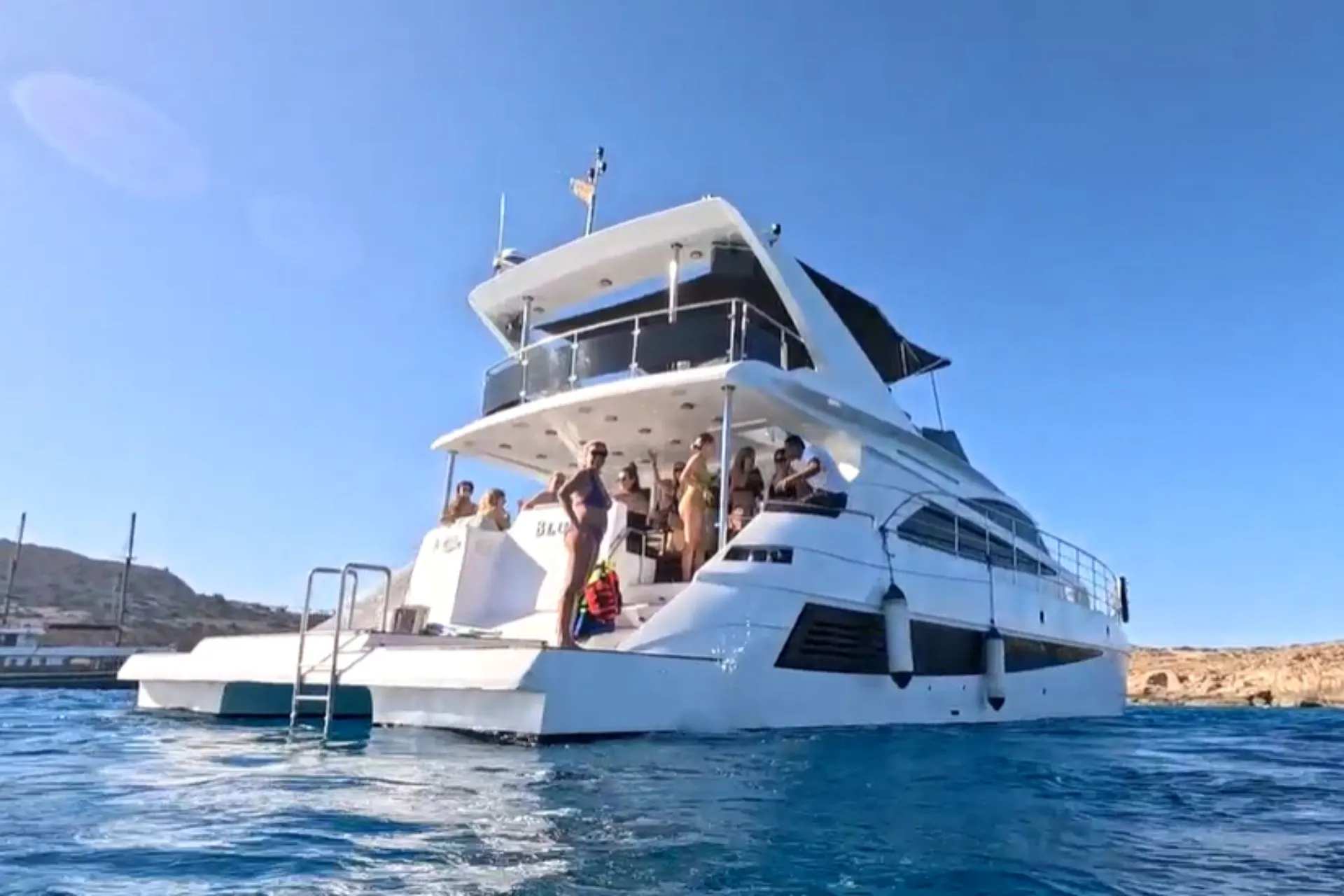 Witte motorjacht met meerdere dekken ligt op helderblauw water, met mensen aan boord en een rotsachtige kustlijn op de achtergrond.