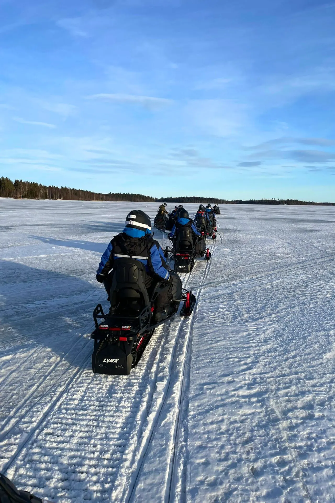 Een stoet sneeuwscooters rijdt achter elkaar door het besneeuwde landschap van Zweden, omringd door ongerepte natuur en een heldere blauwe lucht.