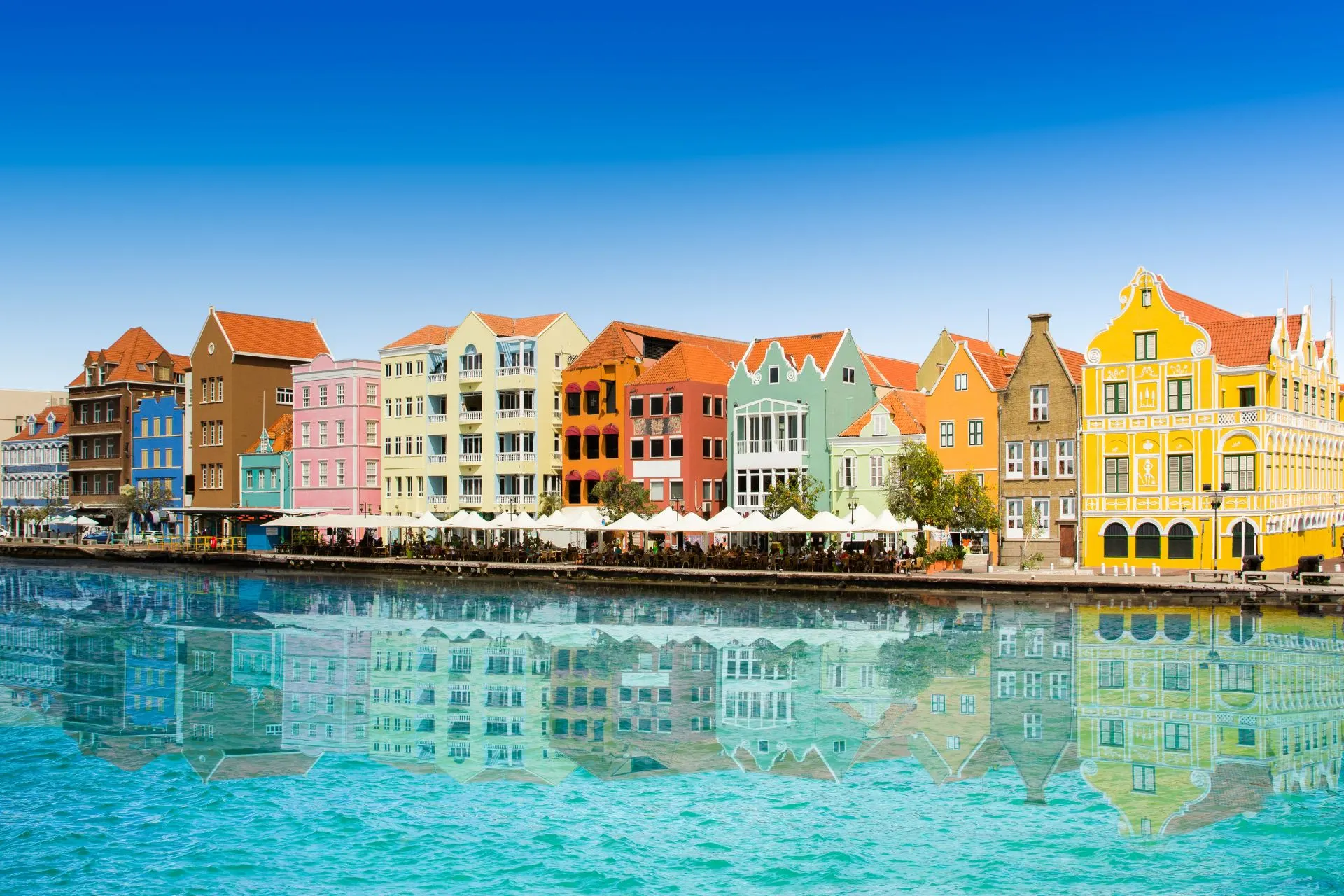 Schilderachtig uitzicht op de kleurrijke gevels van historische panden langs de waterkant in Willemstad, Curaçao. De gebouwen zijn geschilderd in levendige tinten zoals geel, roze, blauw en groen, en weerspiegelen prachtig in het heldere turquoise water van de Caribische Zee. Op de kade staan terrassen met witte parasols, ideaal voor toeristen die genieten van lokale cultuur, architectuur en ontspannen sfeer. Deze iconische locatie is een populaire bestemming voor een vakantie in het Caribisch gebied.