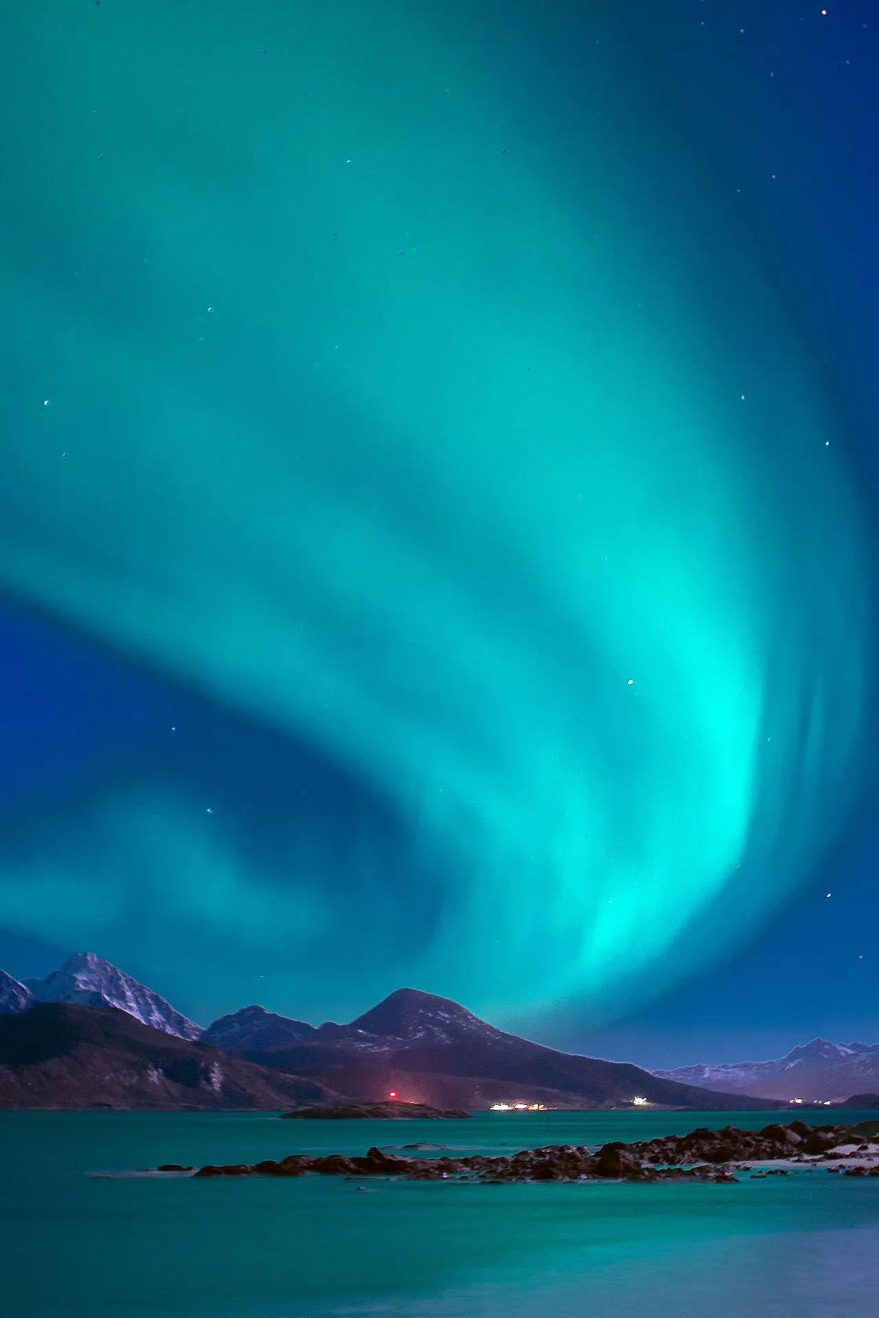 Een levendig groen noorderlicht danst boven een ruige kustlijn met besneeuwde bergen en rustig water, wat een betoverende sfeer creëert die perfect aansluit bij de unieke natuurbeleving van een expeditiecruise in noordelijke gebieden. De reflectie van het licht op zee en de afgelegen ligging benadrukken de exclusiviteit van reizen met kleine expeditieschepen tijdens avontuurlijke poolreizen.