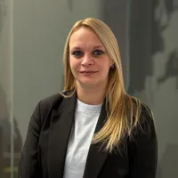 Portret van een vrouw met lang blond haar, gekleed in een zwarte blazer en een wit shirt, staand voor een neutrale achtergrond met glazen panelen. Nicky Coppens