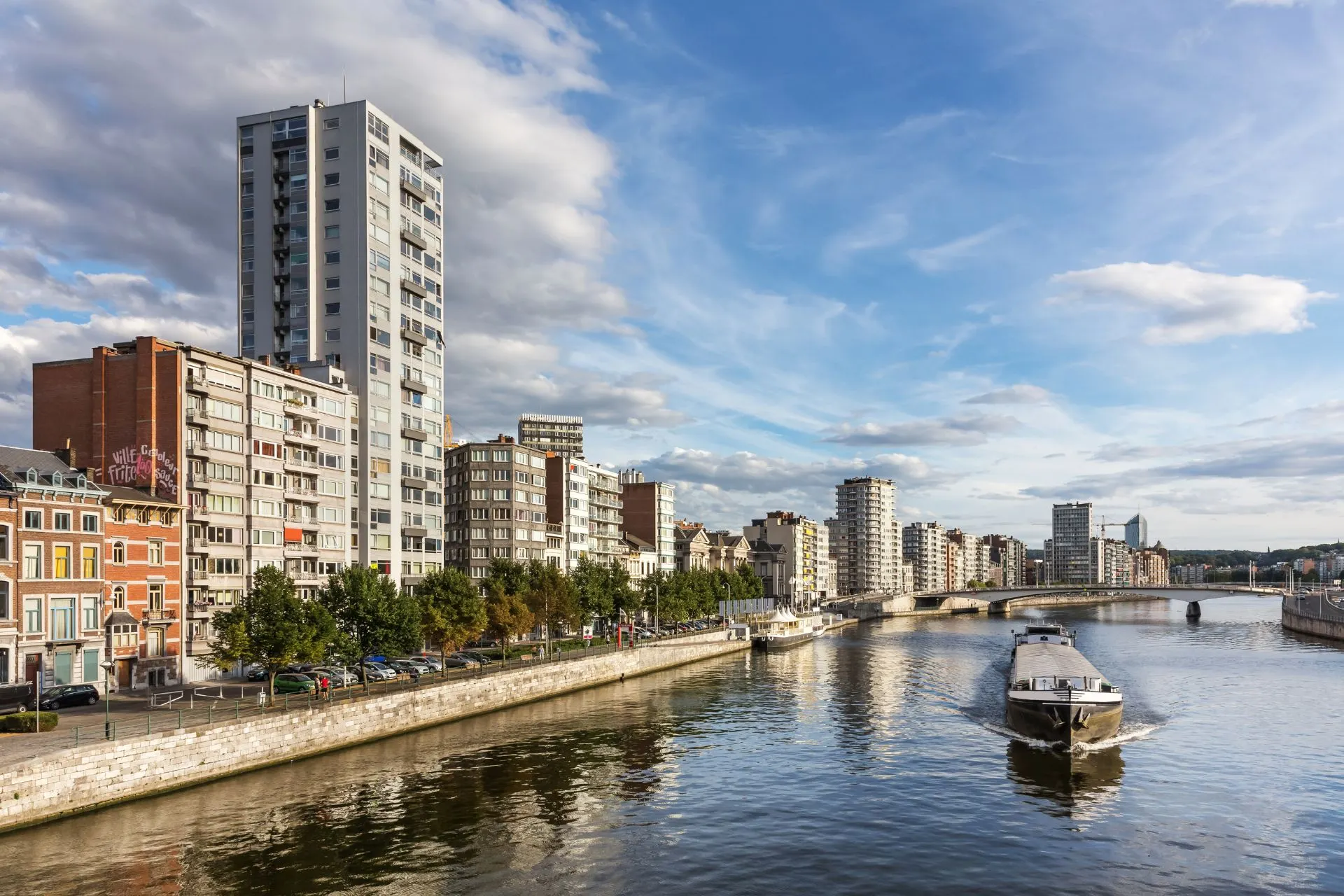 Uitzicht op Luik met een boot op het water langs hoge gebouwen, vlak bij de stad waar je goedkope vluchten vanaf Luik kunt boeken. Ontdek bestemmingen vanaf Liege Airport en geniet van een ontspannen vertrek vanaf luchthaven Luik met voordelige vluchten vanaf Luik Airport.