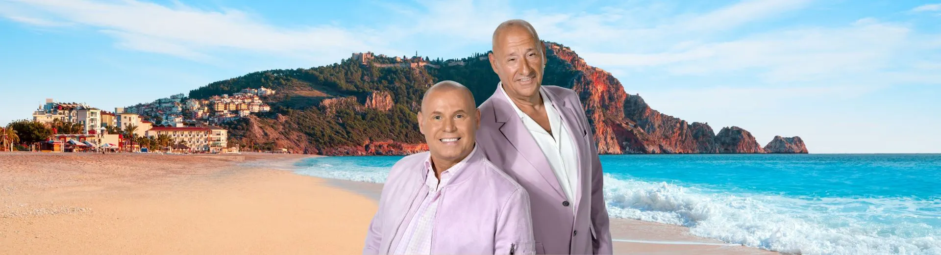 Antonio en Frans staan lachend op een zonnig strand met helderblauwe lucht en zee op de achtergrond. Op de heuvels achter hen liggen kleurrijke appartementen en hotels. Het beeld straalt vakantie, ontspanning en zomerplezier uit.