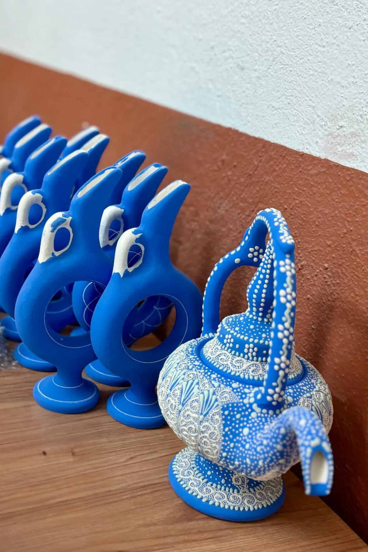 Rij van felblauwe keramische kannen met een opvallend modern design, naast een gedetailleerd beschilderde traditionele waterkan in wit en blauw, geplaatst op een houten oppervlak tegen een rood-witte muur. De kunstige patronen op de grote kan tonen fijne stippen en lijnen, typerend voor Turkse ambachtelijke keramiek uit Avanos, Cappadocië. Deze combinatie van hedendaagse en klassieke stijlen benadrukt de rijke cultuur en vakmanschap van de regio, populair bij toeristen en liefhebbers van handgemaakte kunst.