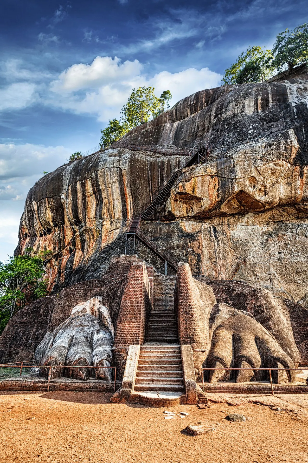 De iconische ingang van de Sigiriya-rots in Sri Lanka, met indrukwekkende uitgehouwen leeuwenpoten aan de voet van de trap die naar de top van de rots leidt. De stenen trap en metalen trappen slingeren omhoog langs de steile, roodbruine rotswand. Deze historische plek, ook wel de Leeuwenrots genoemd, was ooit een koninklijk fort en biedt vandaag de dag een fascinerende blik op de oude koninkrijken van Sri Lanka.
