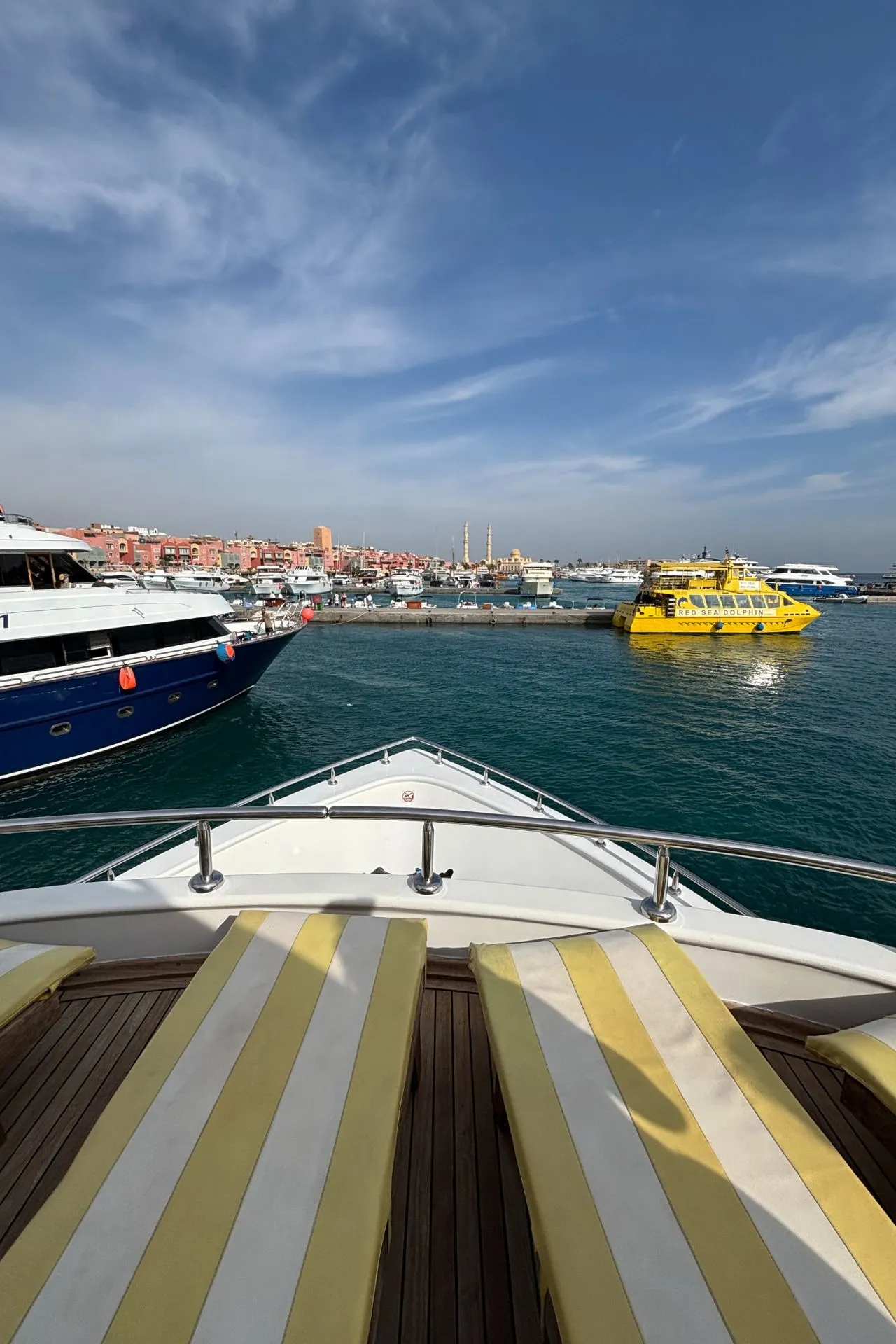 Uitzicht vanaf het dek van een luxe boot met twee geel-wit gestreepte ligbedden op de voorgrond, gericht op een levendige haven. In het heldere turquoise water liggen meerdere boten, waaronder een opvallende gele veerboot en een grote blauwe jacht. Op de achtergrond zijn kleurrijke gebouwen en minaretten zichtbaar onder een blauwe lucht met lichte wolken. Dit beeld benadrukt ontspanning en luxe tijdens een boottocht, ideaal voor promotie van havenexcursies, cruises en vakanties aan de Rode Zee in Egypte.