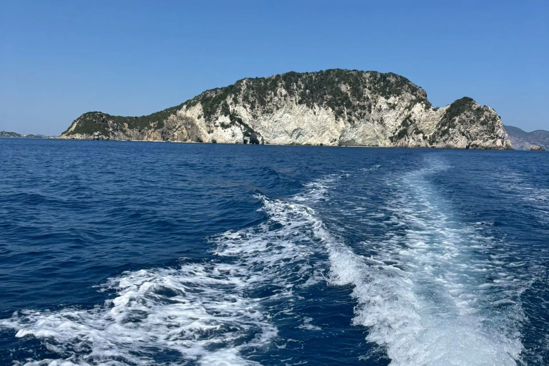 Uitzicht vanaf een boot op helderblauw water met schuimspoor en een rotsachtig eiland aan de horizon bij Zakynthos.