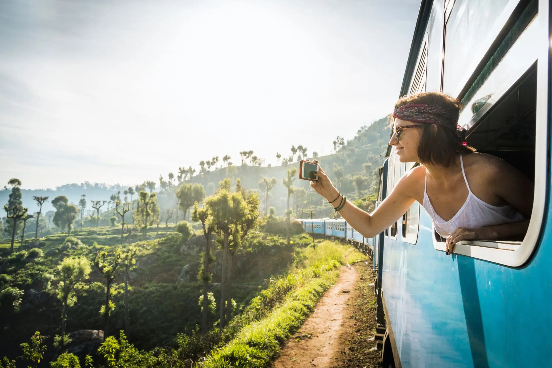 Vrouw hangt uit het raam van een blauwe trein en maakt een foto van het tropische, heuvelachtige landschap in Sri Lanka.