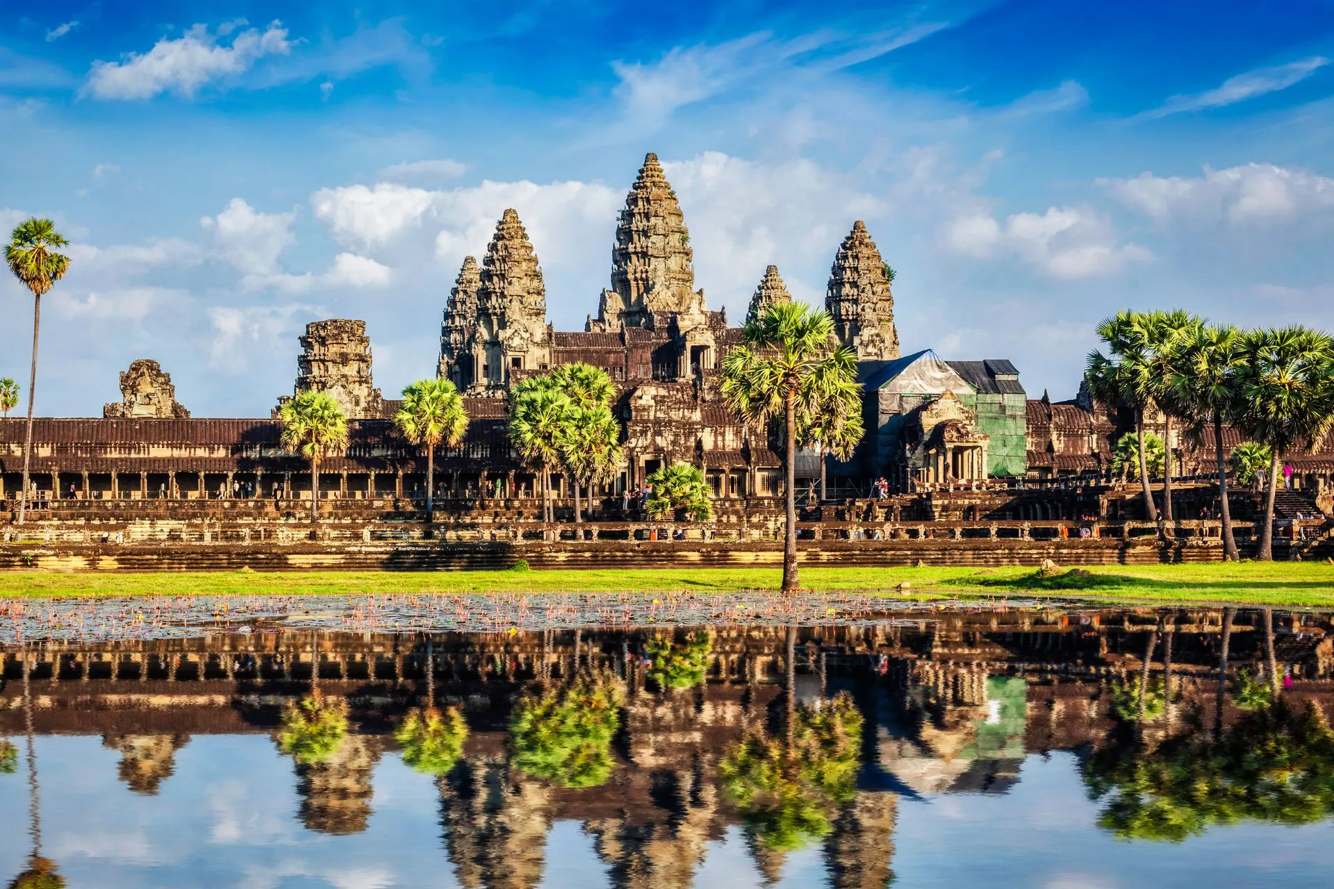 Uitzicht op een groot tempelcomplex met torens en palmbomen, weerspiegeld in een vijver onder een heldere blauwe lucht.