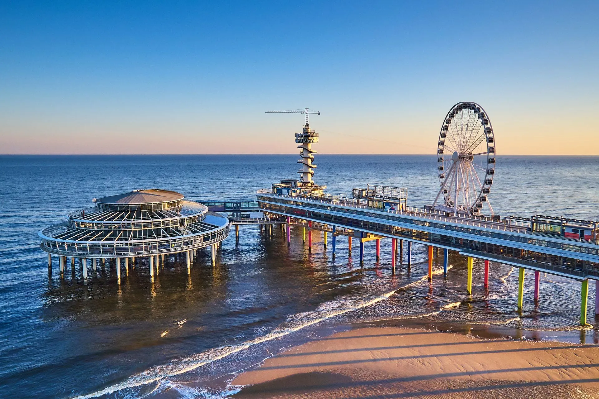 De pier van Scheveningen met het reuzenrad en de zee op de achtergrond, bij zonsondergang met warme kleuren en rustige branding.

