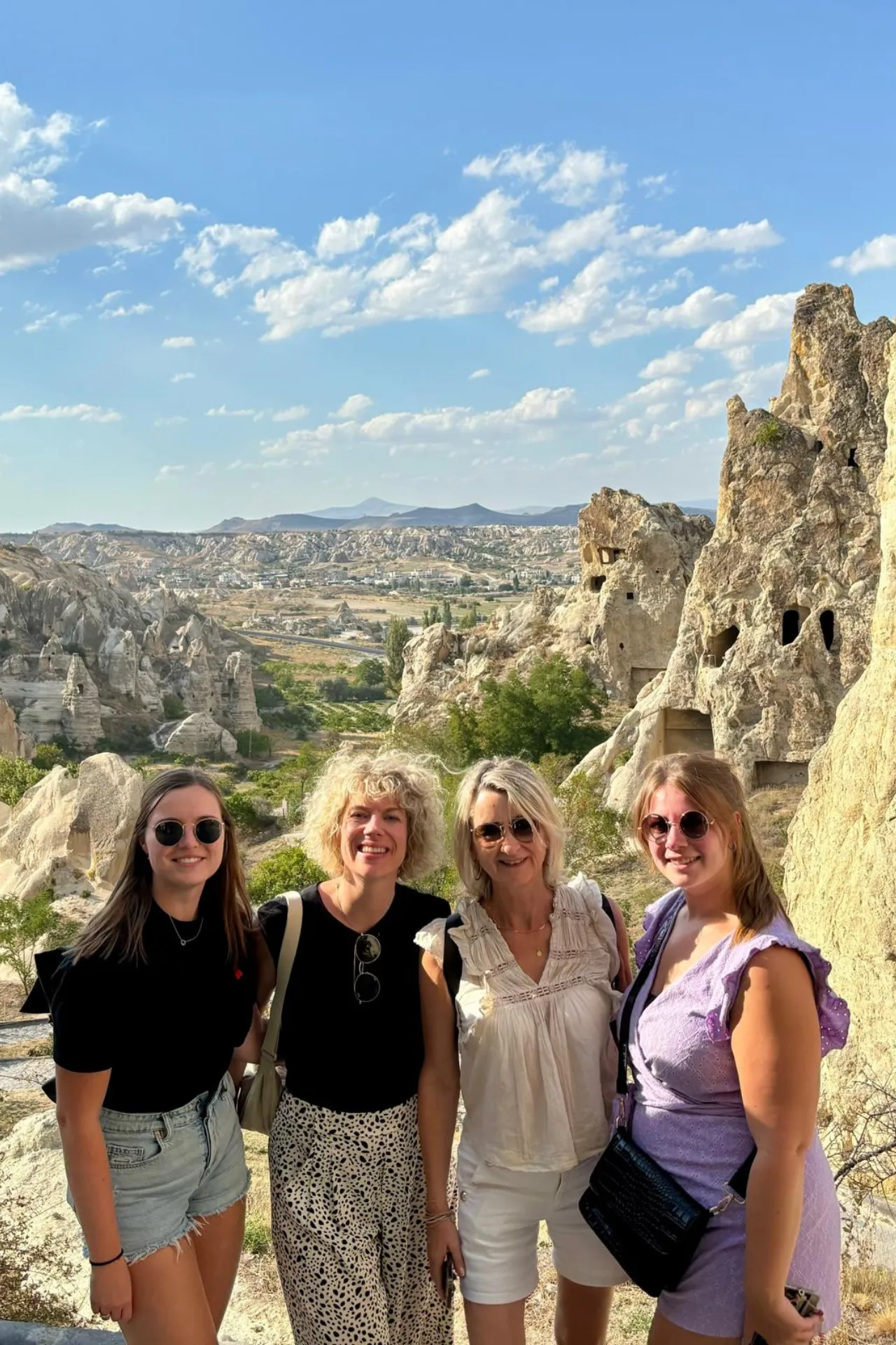 Vier personen poseren voor een foto in het indrukwekkende rotslandschap van Cappadocië, Turkije. Op de achtergrond zijn grillige rotsformaties en uitgehouwen grotten zichtbaar, typerend voor deze historische regio die bekendstaat om zijn unieke geologie en culturele erfgoed. De heldere blauwe lucht met verspreide wolken benadrukt het zonnige weer, terwijl de natuurlijke tinten van het gesteente een prachtig contrast vormen. Deze locatie is een populaire bestemming voor toeristen en fotografen die op zoek zijn naar bijzondere bezienswaardigheden in Anatolië.