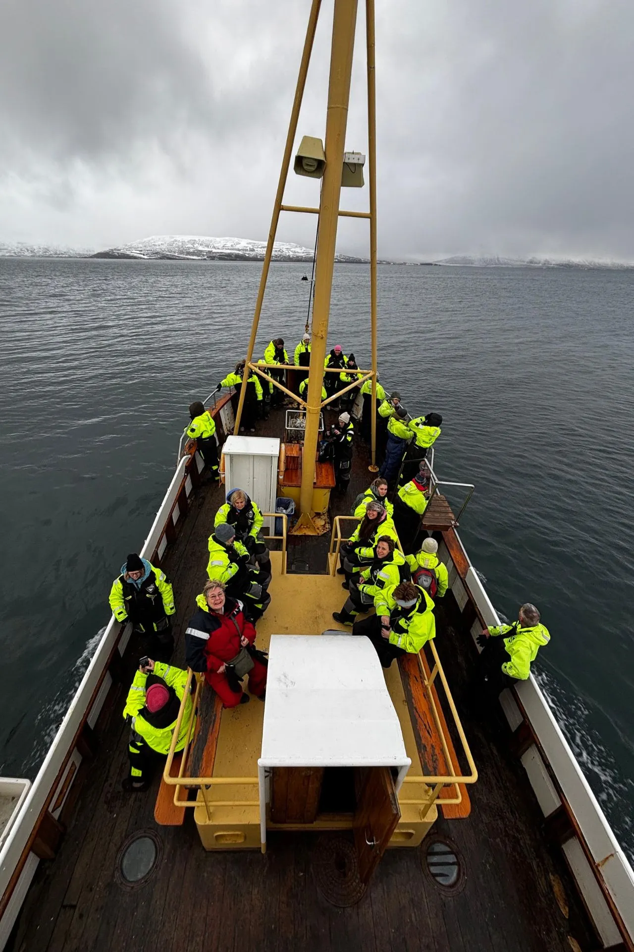 Groep reizigers in winterkleding op een houten walvissafariboot in Noord‑IJsland, varend door een fjord.
