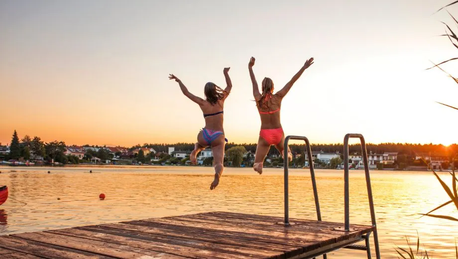 Twee jonge vrouwen springen hand in hand in een meer vanaf een houten steiger, precies op het moment dat de zon ondergaat. Ze dragen beiden een kleurrijke bikini en zijn vastgelegd in de lucht met de armen omhoog in een uitbundige en vrolijke pose. Het meer ligt in een rustige vakantiebestemming omringd door vakantiehuizen, bomen en een kalme omgeving. De lucht is oranje en goudkleurig door de ondergaande zon, wat de hele scène een warme zomerse gloed geeft. Aan de linkerkant is een rode kano zichtbaar in het water. Deze levendige vakantieafbeelding straalt vrijheid, ontspanning en avontuur uit – perfect voor marketing rondom actieve vakanties, zomeraanbiedingen, jongerenreizen, of gezinsvakanties aan het water. Ideaal voor vakantie boeken naar bestemmingen met meren, zwemgelegenheden of natuurcampings.