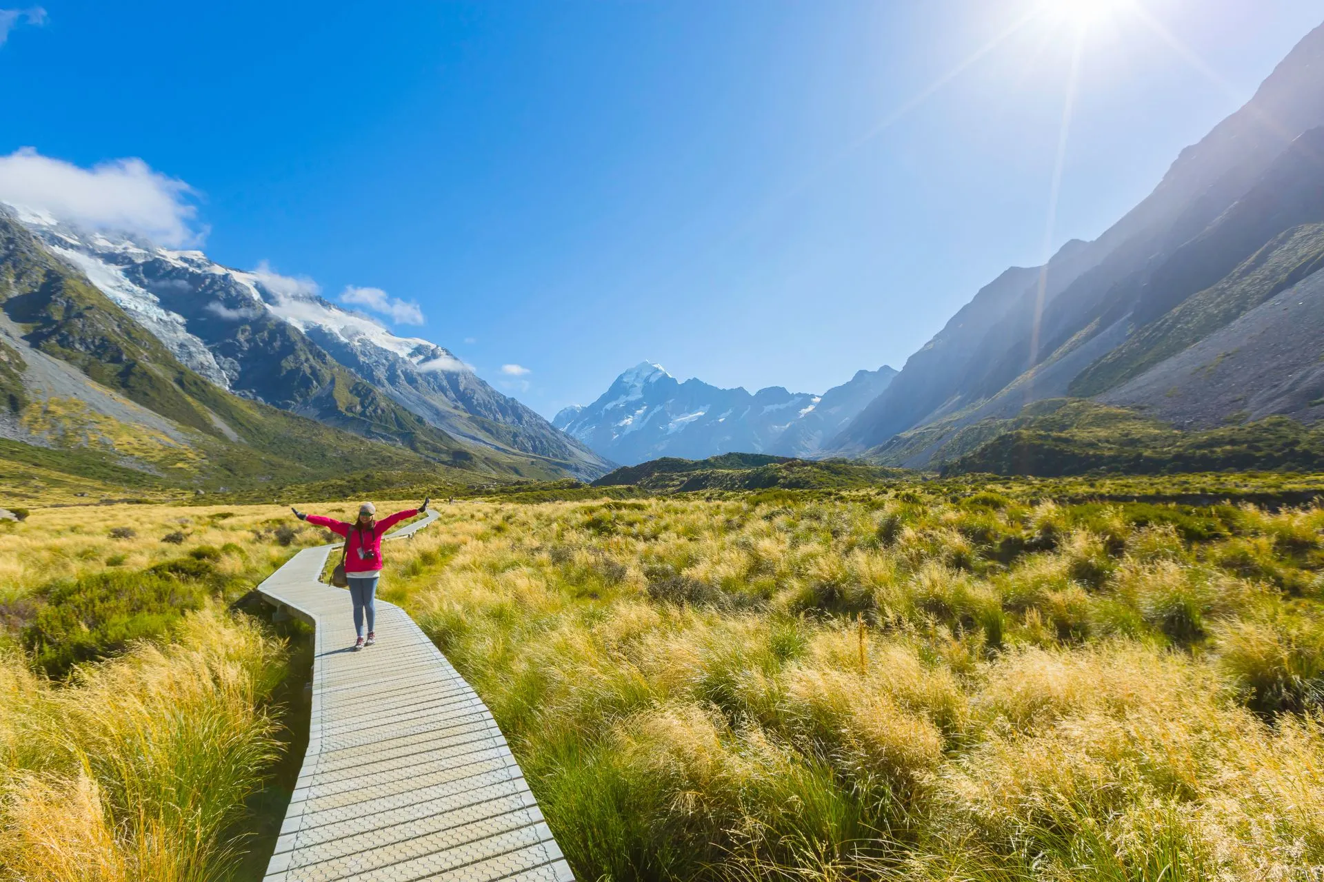 Indrukwekkend uitzicht op Mount Cook in Nieuw-Zeeland, met een houten wandelpad dat door een uitgestrekt veld van goudkleurige graslanden slingert richting de imposante besneeuwde bergtoppen. De heldere blauwe lucht en zonnestralen verlichten het berglandschap, wat een serene en avontuurlijke sfeer creëert. Perfecte bestemming voor wandeltochten, fotografie en natuurbeleving. Ideaal voor reizigers die op zoek zijn naar avontuur, rust en het ontdekken van de ongerepte schoonheid van de Zuidelijke Alpen.