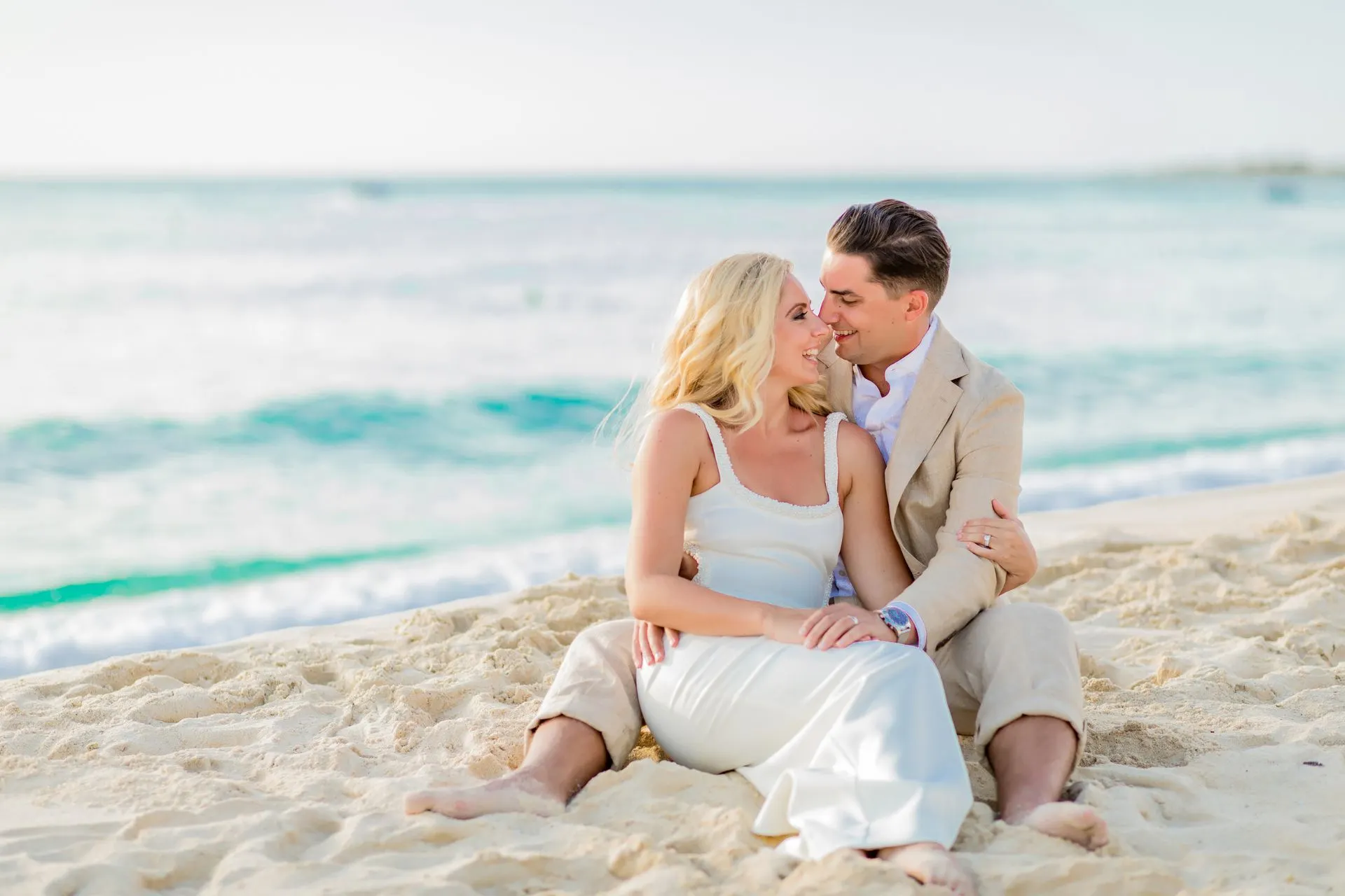 Een romantisch moment van een pasgetrouwd stel zittend op het witte zandstrand van Aruba. De bruid draagt een elegante witte jurk, terwijl de bruidegom een beige pak draagt. Ze lachen naar elkaar en genieten van de serene omgeving met turquoise water en een kalme zee op de achtergrond.
