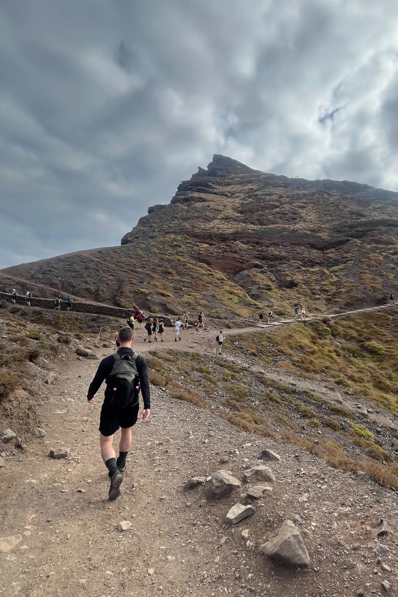 Groep wandelaars op rotsachtig bergpad richting een piek op Madeira onder een dramatische wolkenlucht.