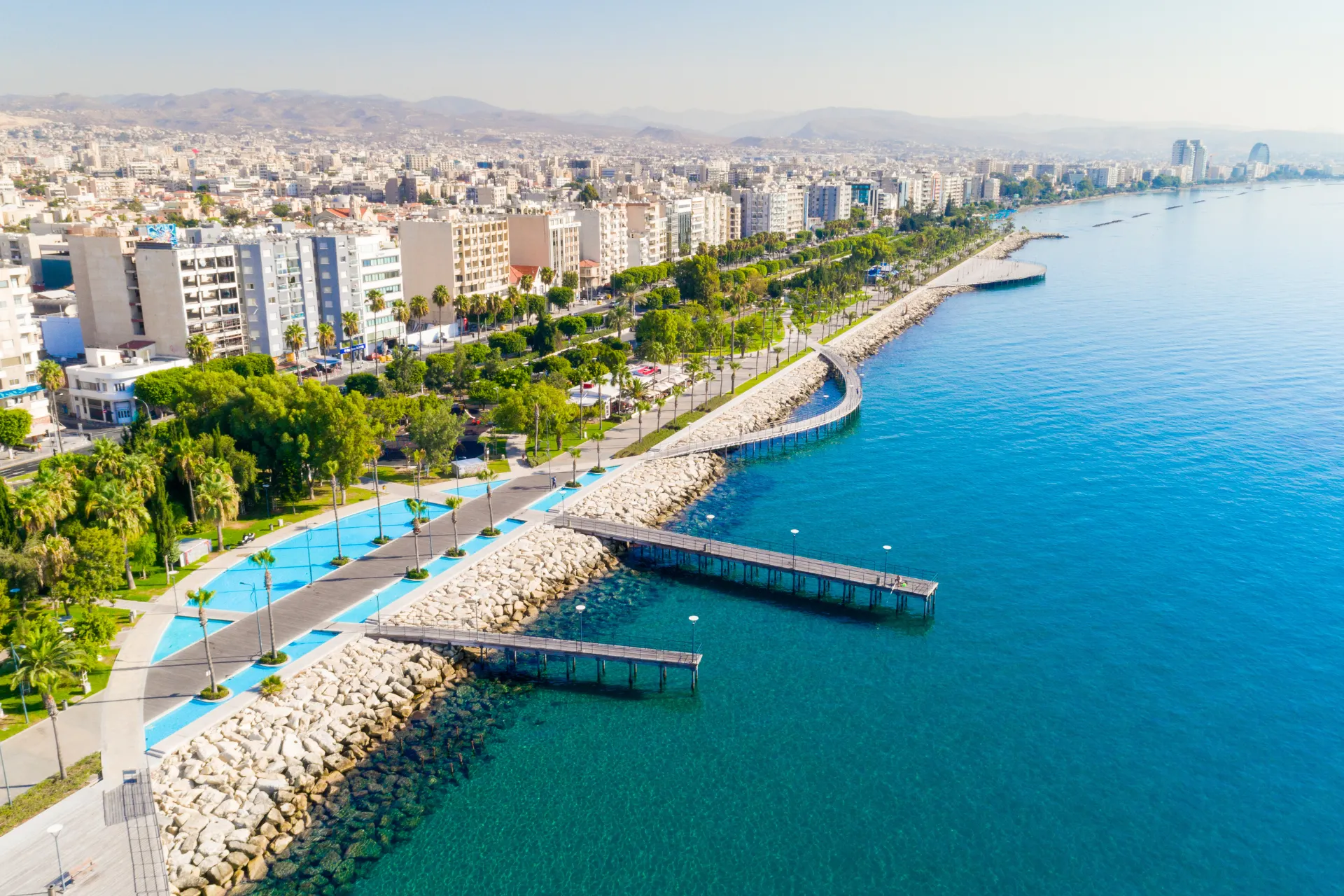 "Panoramisch uitzicht op een moderne boulevard langs de kust met helderblauw water, palmbomen en een lange rij gebouwen tegen een bergachtig landschap. Perfecte bestemming voor een mediterrane vakantie, ideaal voor zon, zee, cultuur en ontspanning. Kristalheldere zee, groene parken en levendige stadssfeer maken deze plek tot een paradijs voor reizigers die houden van fotografie, gastronomie en unieke ervaringen."