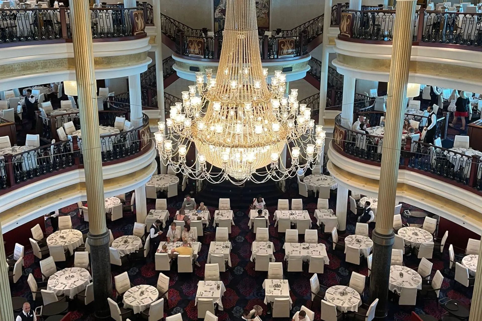 Luxe hoofdrestaurant van de Independence of the Seas met ronde en rechthoekige tafels, meerdere verdiepingen en een indrukwekkende kristallen kroonluchter in het midden.