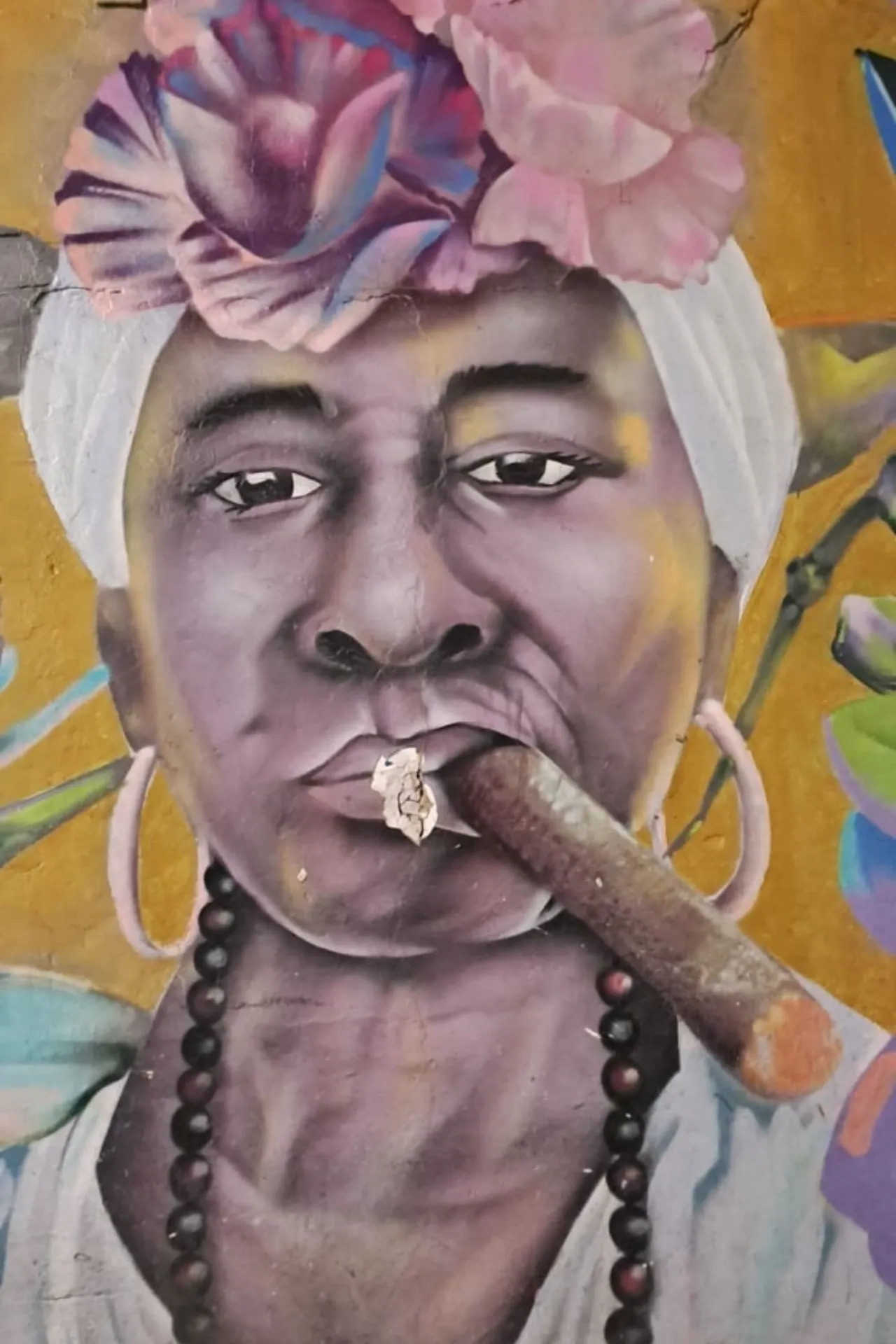 Artistieke muurschildering van een persoon met bloemen op het hoofd en kralenketting, tegen een gele achtergrond met plantenmotieven.