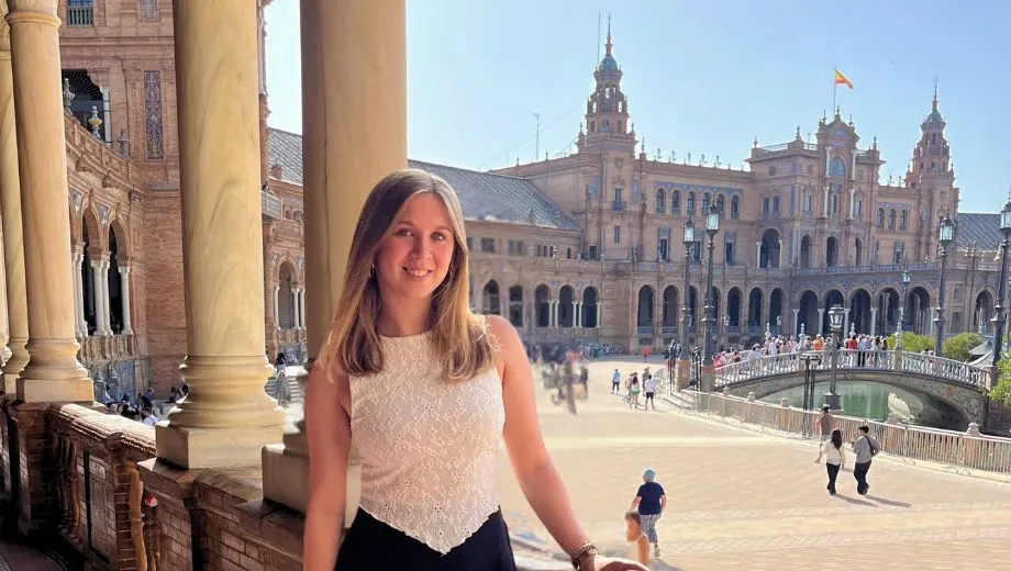 Marketeer Martje poseert glimlachend op het balkon van Plaza de España in Sevilla. Ze draagt een witte kanten top en zwarte shorts. Op de achtergrond zijn de halfronde galerijen, torens en het kanaal van het iconische plein duidelijk zichtbaar, met mensen die over de brug wandelen.