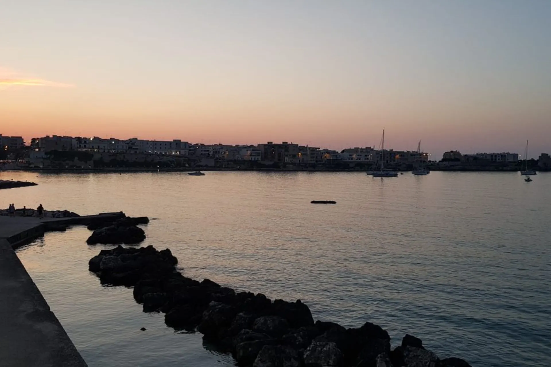 Een sfeervol uitzicht op de kust van Puglia tijdens zonsondergang, met de zee en de verlichte stad op de achtergrond.