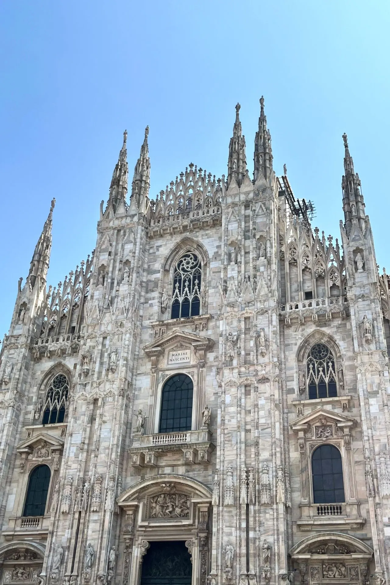 Vooraanzicht van de Duomo di Milano, met zijn kenmerkende spitsbogen, beelden en grote ramen. De lucht is helderblauw en wolkeloos.