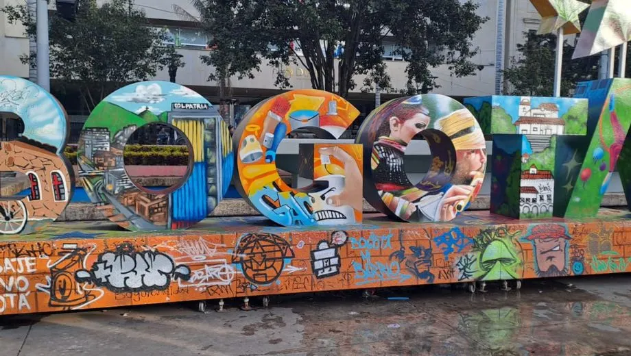 Grote letters die samen “BOGOTÁ” vormen, beschilderd met kleurrijke graffiti en illustraties van stadsgezichten en culturele elementen, geplaatst op een plein.
