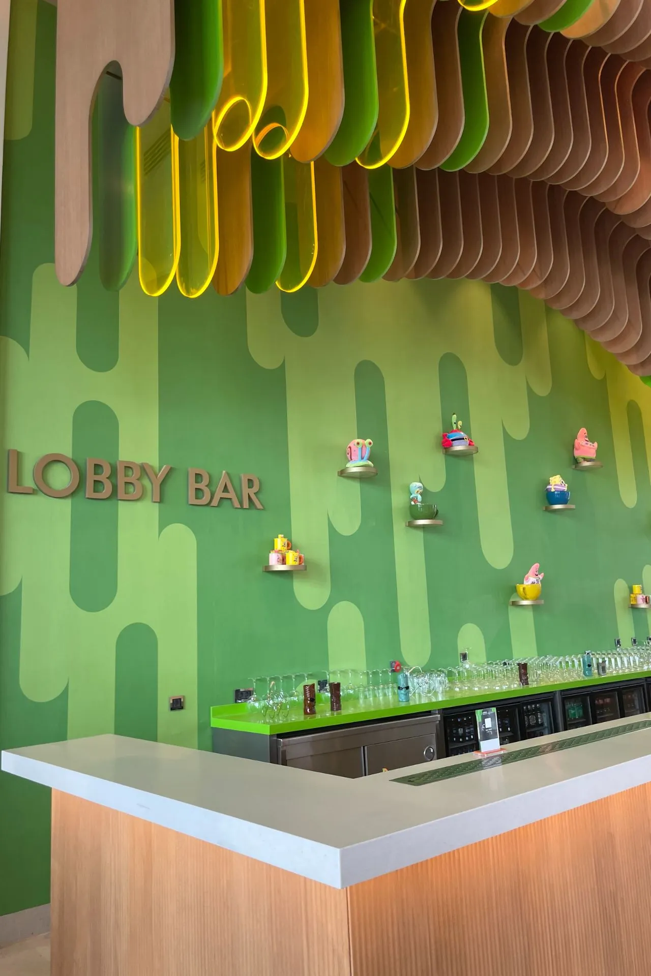Moderne lobbybar met groene wanddecoratie, houten golvende panelen en kleurrijke miniatuurfiguren op de muur.