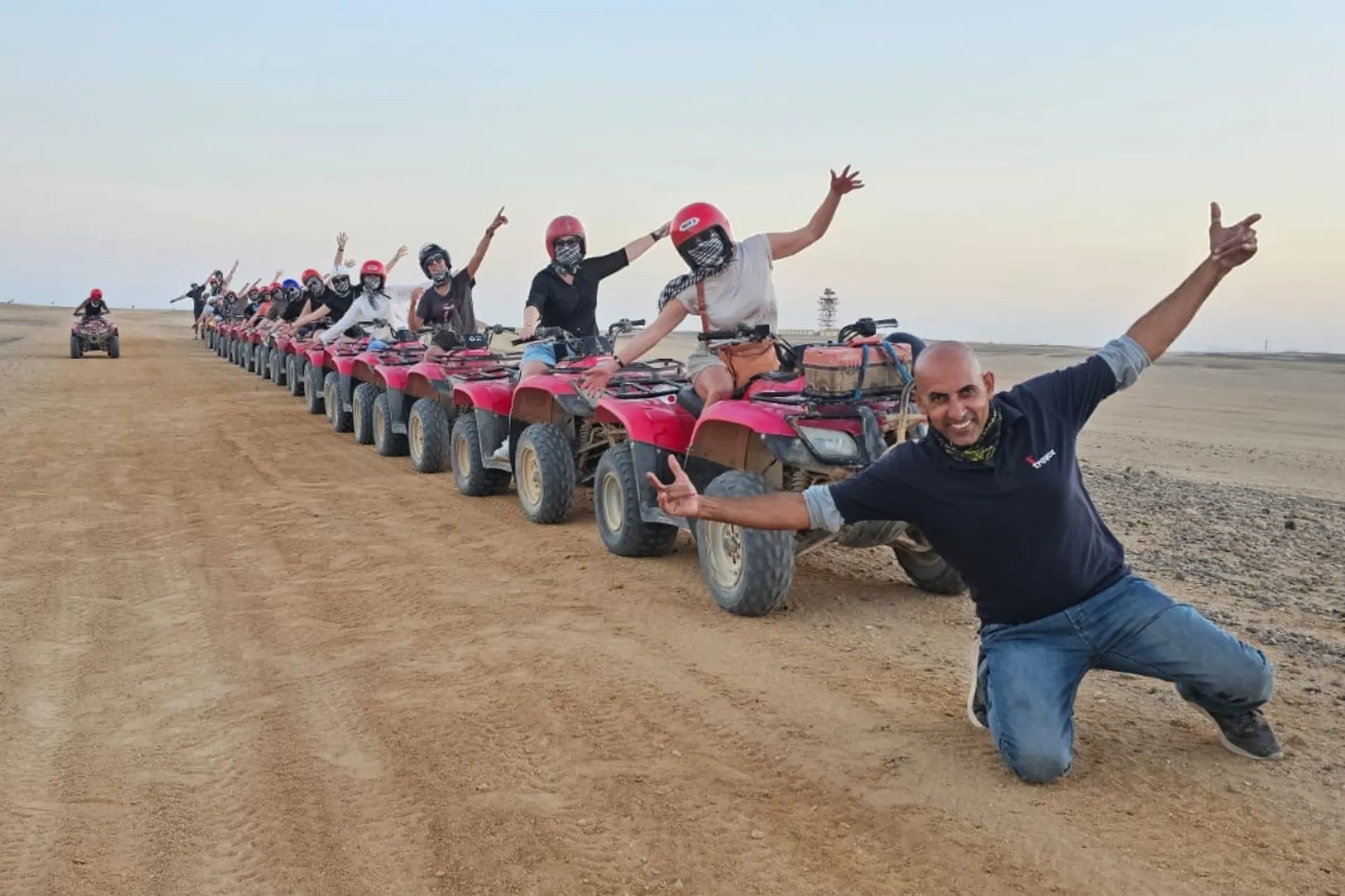 Een lange rij rode quads staat opgesteld op een zandpad in een uitgestrekt woestijnlandschap, klaar voor een avontuurlijke rit. Mensen zitten op de quads met helmen en sjaals ter bescherming tegen stof, terwijl een begeleider vooraan enthousiast poseert met uitgestrekte armen. Op de achtergrond is een vlakke horizon zichtbaar onder een heldere lucht, wat een gevoel van vrijheid en avontuur oproept. Ideaal beeld voor promotie van quadtochten, woestijnexcursies en actieve vakanties in Egypte.