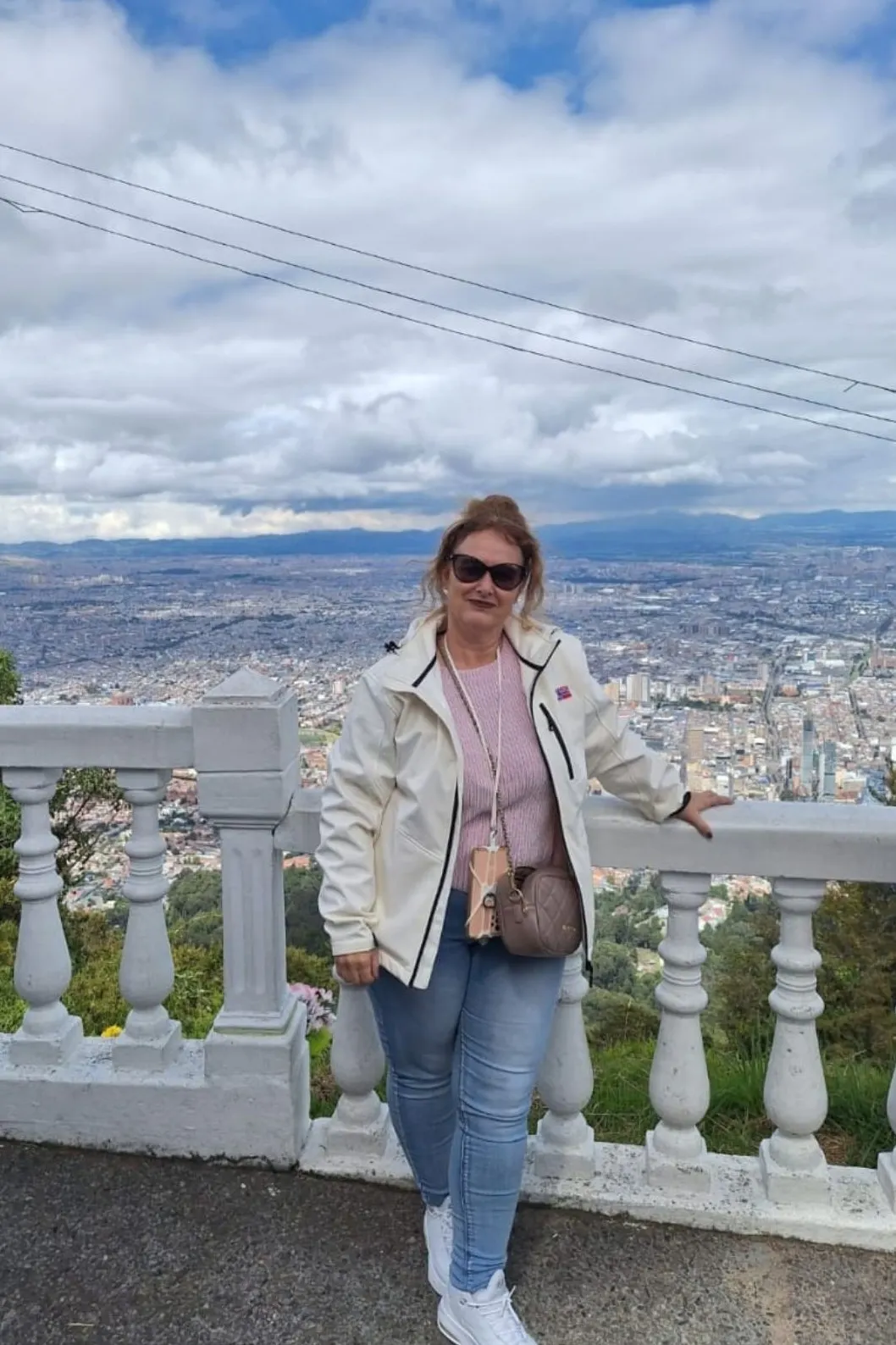 Uitzichtpunt met witte balustrade en panoramisch zicht over een grote stad, bergen en wolken op de achtergrond.