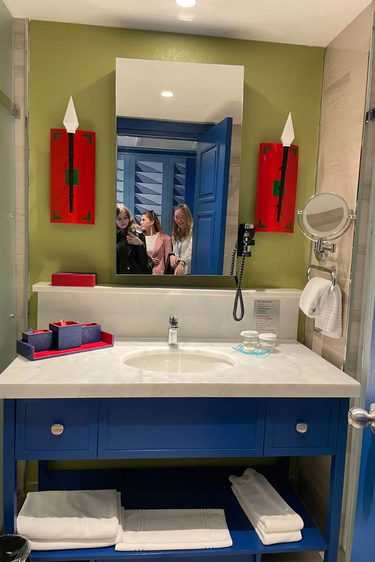  Moderne hotelbadkamer met blauwe kastjes, witte wastafel en spiegel waarin twee personen zichtbaar zijn, met rode decoratieve elementen aan de muur.