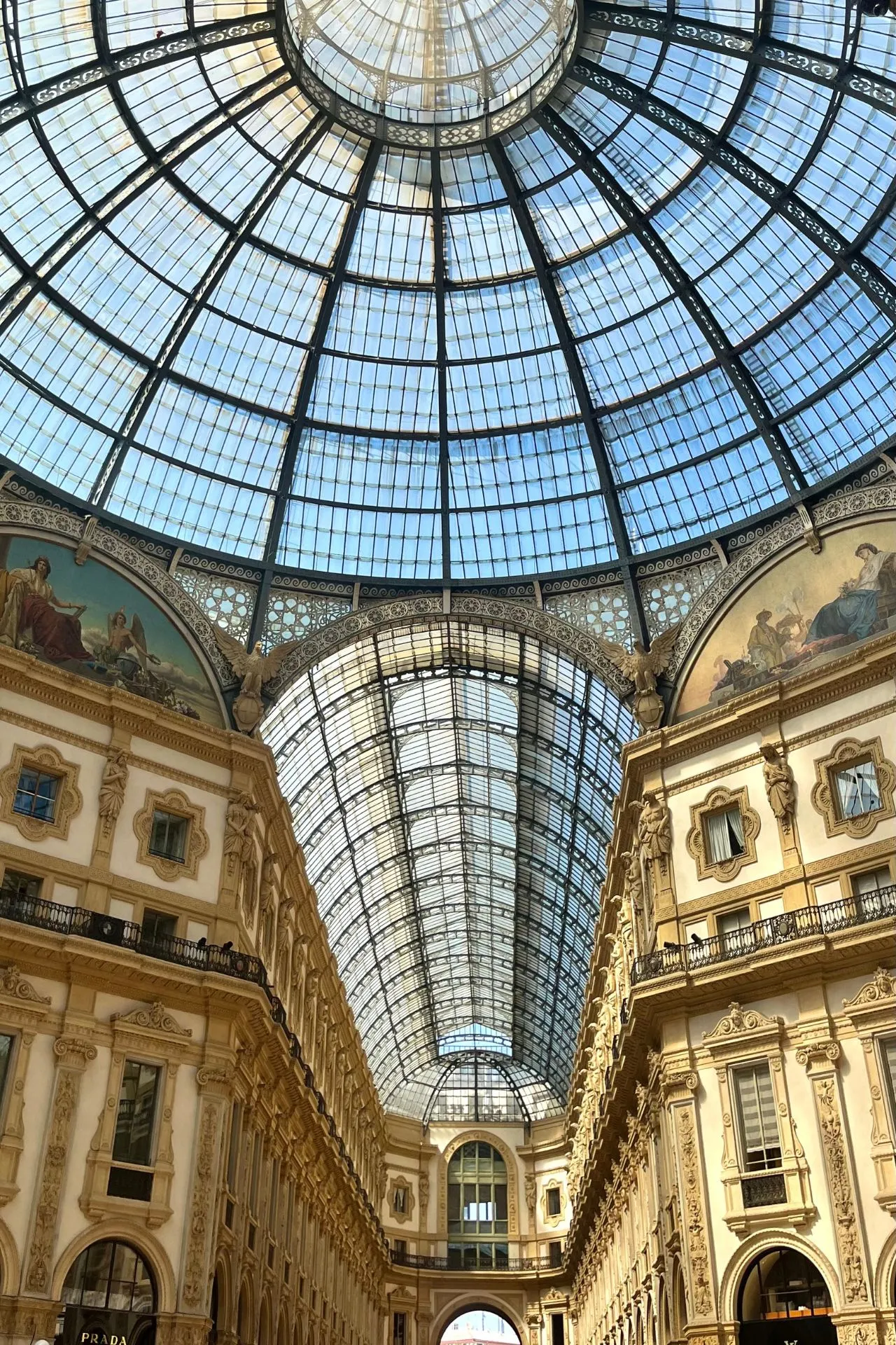 Detail van de glazen overkapping en sierlijke gevels van de Galleria Vittorio Emanuele II, met sculpturen en decoratieve elementen langs de wand en het balkon.

