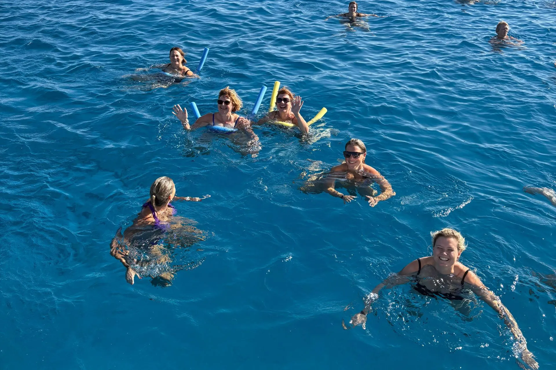 Meerdere mensen zwemmen in helderblauw water, sommigen met drijvende zwemstokken, onder een zonnige blauwe lucht.