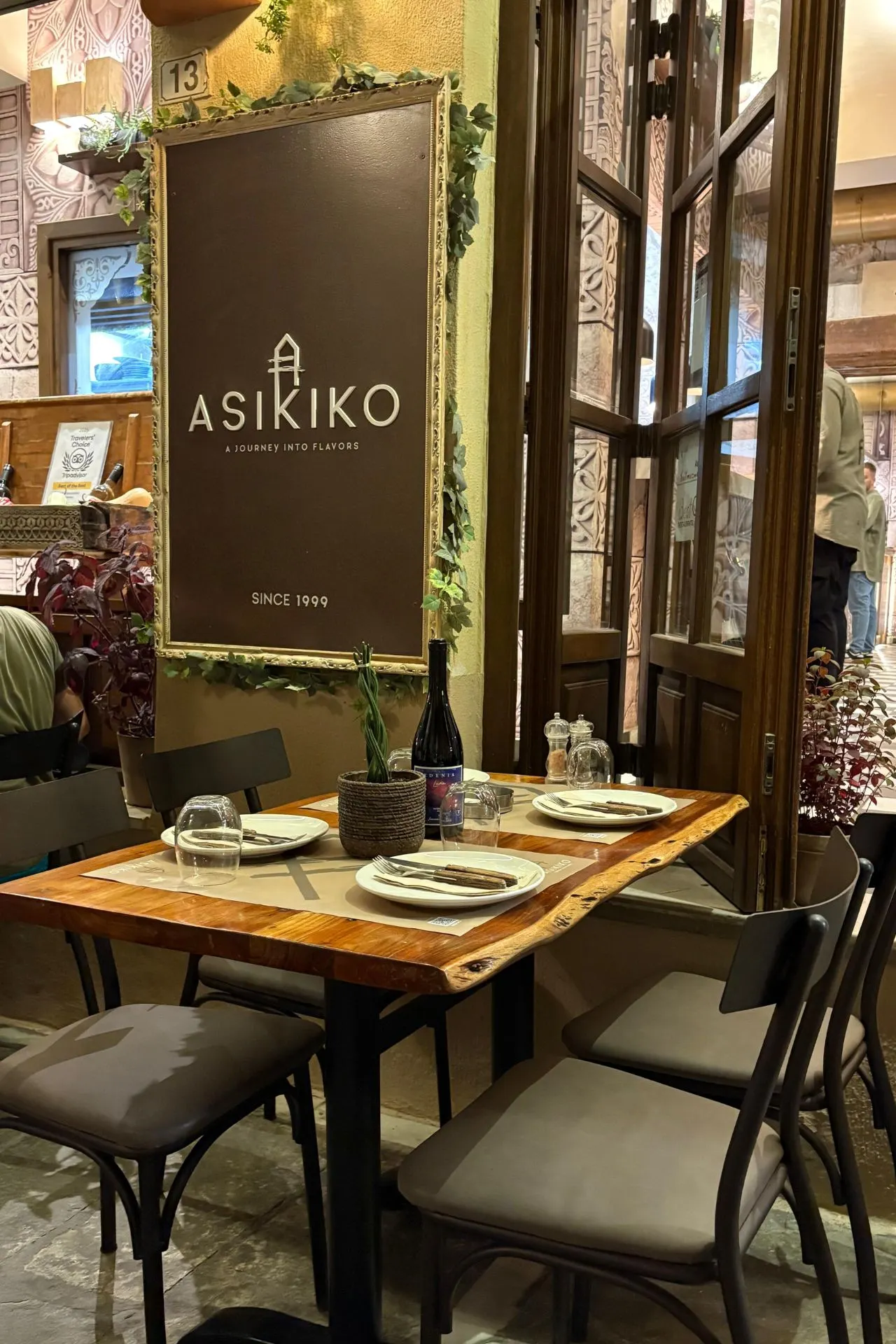 Ingang van restaurant Asikiko in Rethymnon met houten tafel, gedekte borden en wijnfles, sfeervol verlicht in de avond.