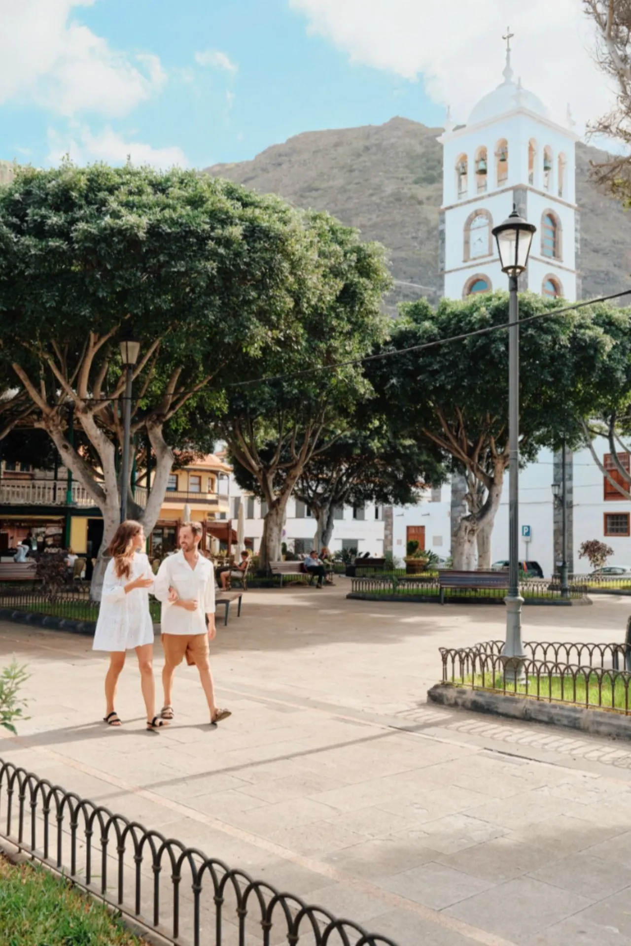 Dorpsplein in Noord‑Tenerife met historische kerk, schaduwrijke bomen en bergen op de achtergrond. In de dorpen van Noord‑Tenerife proef je het lokale leven. Rustige pleinen, historisch erfgoed en omringende bergen zorgen voor een ontspannen sfeer, ver weg van de grote badplaatsen in het zuiden van het eiland. 
