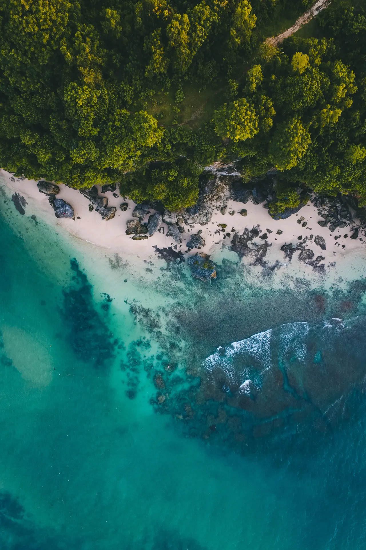 Luchtfoto van bovenaf van een helder blauw strand in Indonesië, omringd door groene bomen en tropische natuur.