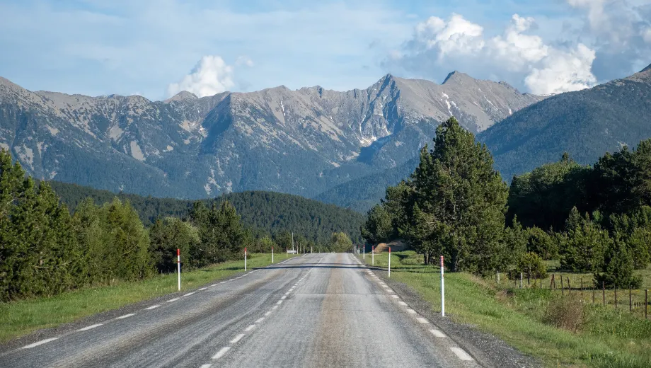 Uitgestrekte berglandschap in Frankrijk met een rechte, rustige weg die door groene bossen en velden slingert richting imposante grijze bergtoppen. De helderblauwe lucht met enkele wolken benadrukt de natuurlijke schoonheid van deze route, ideaal voor een autovakantie of roadtrip door Frankrijk. Deze schilderachtige omgeving biedt een perfecte combinatie van avontuur en ontspanning, met panoramische uitzichten en ongerepte natuur die uitnodigen tot ontdekken en genieten van het Franse platteland.