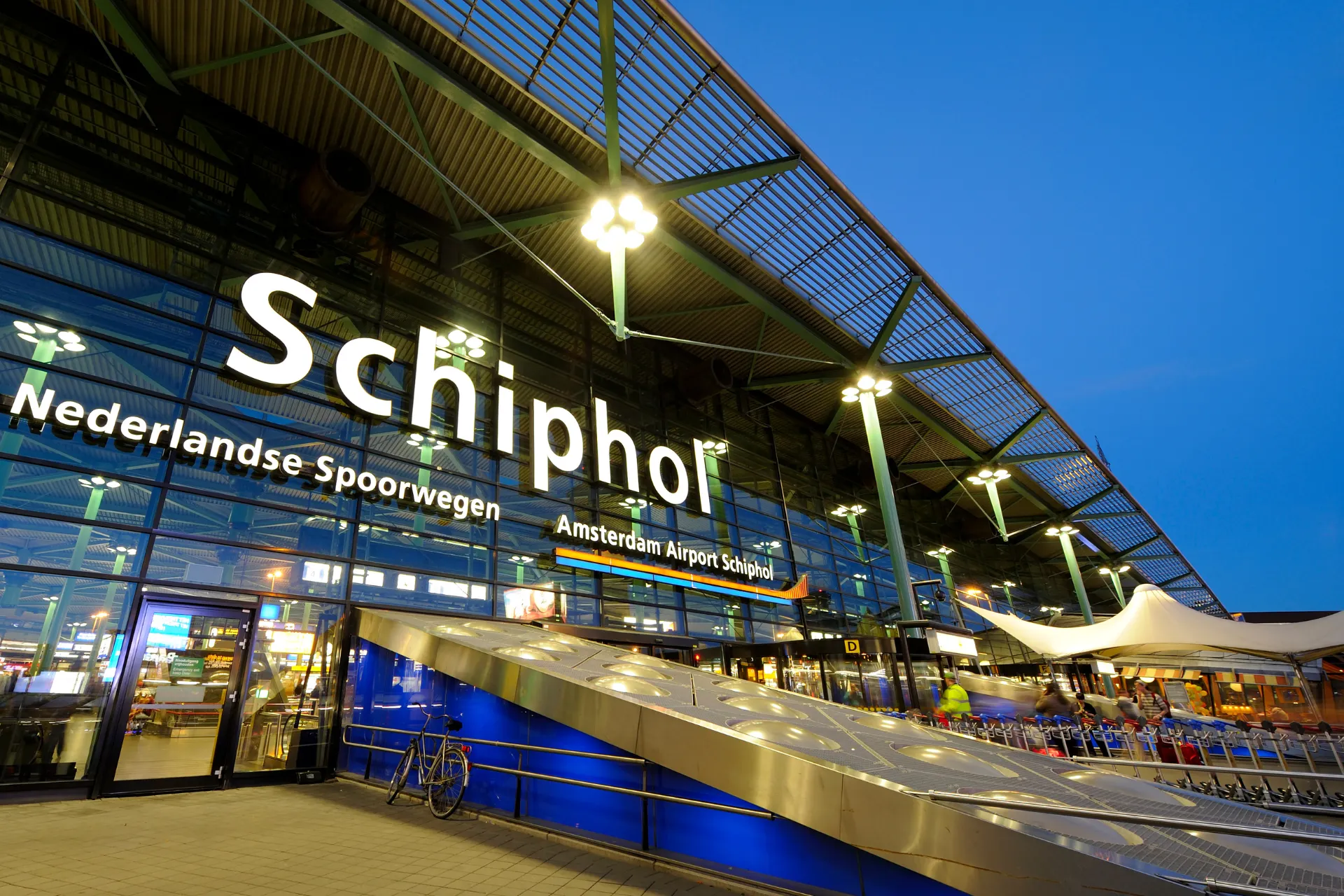 De voorkant van Schiphol bij schemering, waar reizigers zich voorbereiden op vliegen vanaf Amsterdam. Ervaar het gemak van de grootste luchthaven van Nederland.