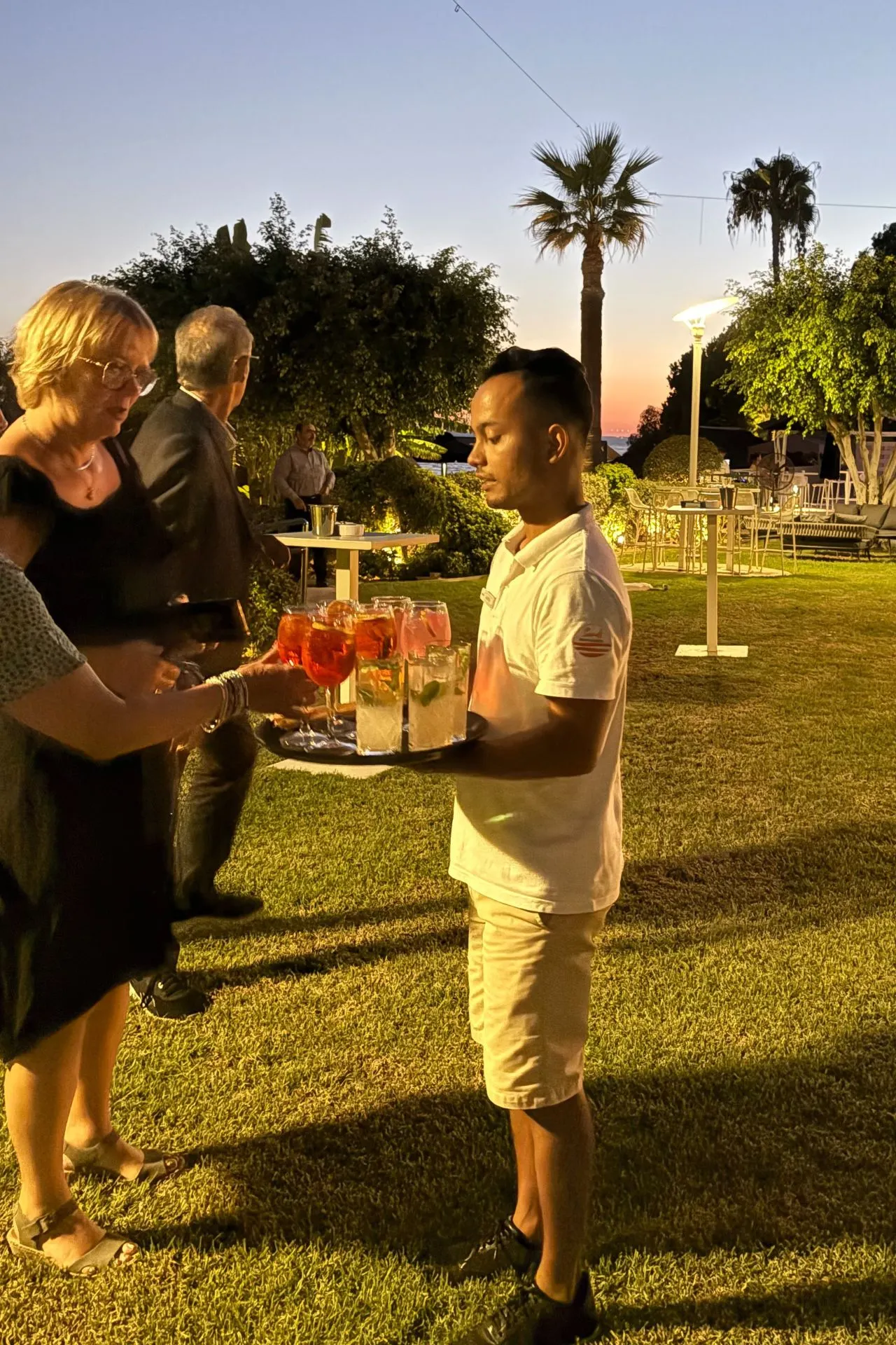 Ober met een dienblad vol cocktails en drankjes in een verlichte tuin met palmbomen, terwijl gasten staan en praten bij hoge tafels.