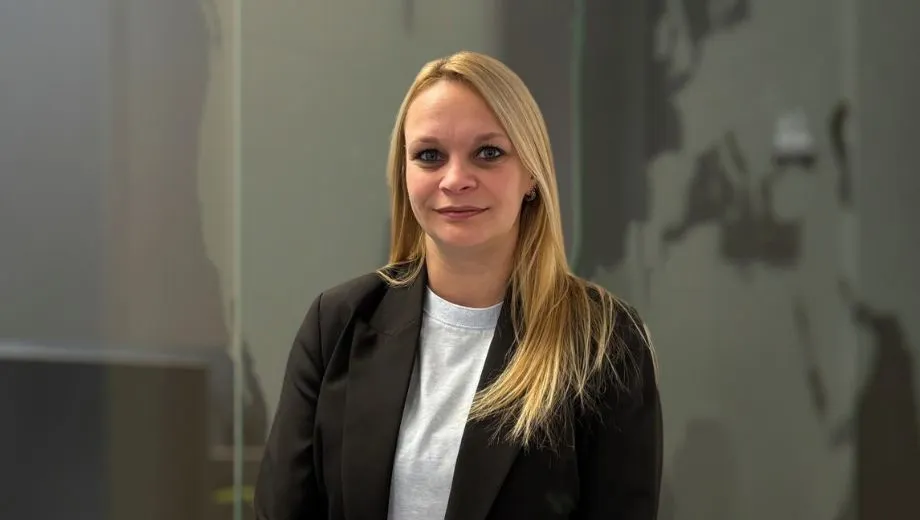 Portret van een vrouw met lang blond haar, gekleed in een zwarte blazer en een wit shirt, staand voor een neutrale achtergrond met glazen panelen. Nicky Coppens