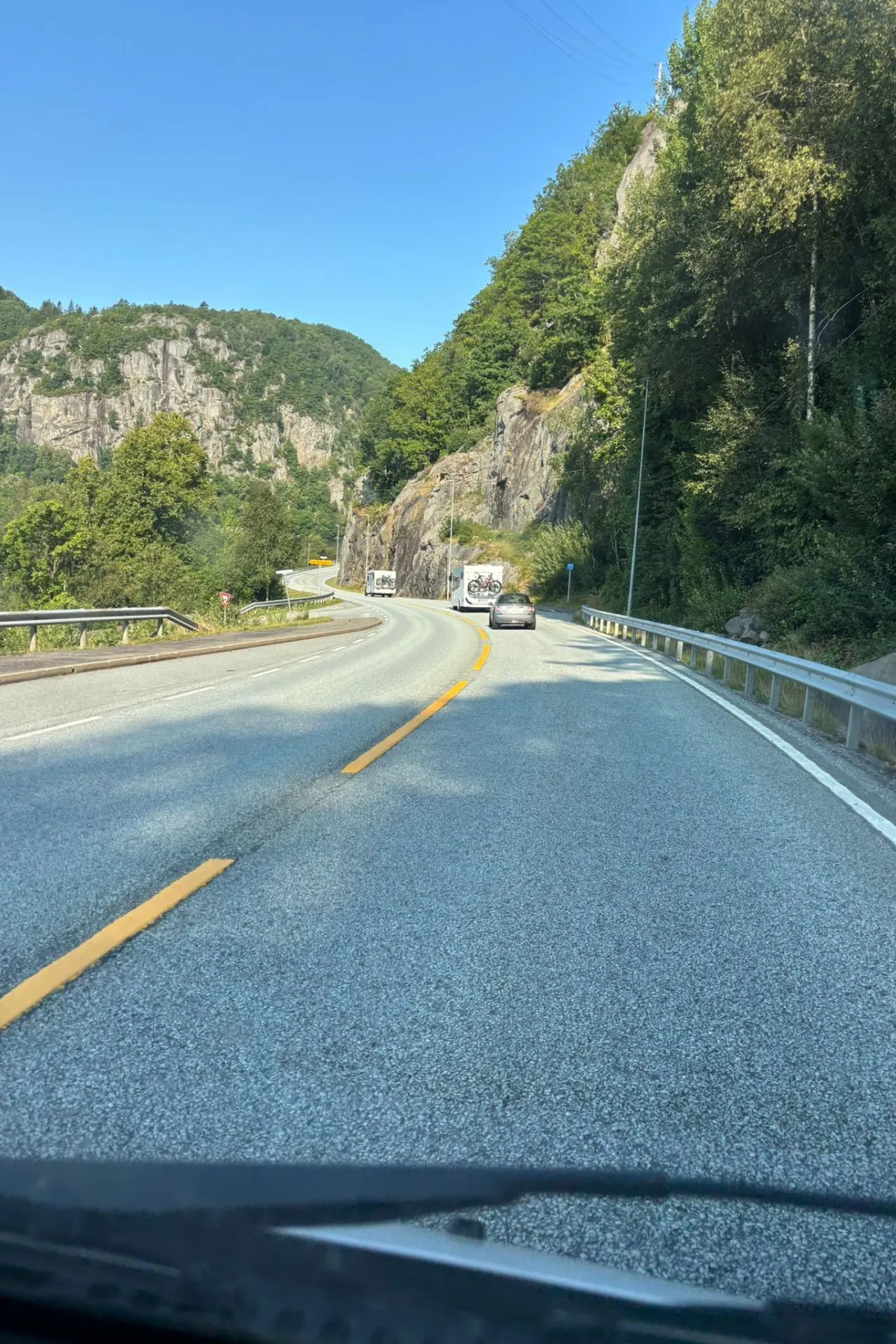 Bochtige bergweg met dubbele gele middenstreep, omgeven door groene bomen en rotswanden onder een heldere blauwe lucht. Een auto rijdt vooraan, gevolgd door campers in de verte.