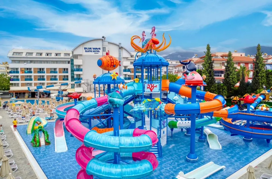 Deze afbeelding toont een kleurrijk waterpark bij een hotel, perfect voor een zonvakantie Verasole of een gezinsvriendelijke Verasole vakantie. Op de voorgrond zie je verschillende blauwe en roze glijbanen, vrolijke waterspeeltoestellen en fonteinen, ideaal voor kinderen en gezinnen die kiezen voor een pakketreis Verasole. Op de achtergrond staat het hotel “Blue Wave Suite Hotel”, omringd door ligbedden en parasols, wat direct een ontspannen vakantiesfeer oproept. De lucht is helderblauw en de omgeving straalt plezier en comfort uit. Deze setting benadrukt het gemak en de veelzijdigheid van Verasole vakanties: alles is tot in de puntjes geregeld, van accommodatie tot waterpret. Of je nu kiest voor een all-inclusive resort of een hotel met aquapark, bij Verasole vind je altijd de perfecte zonvakantie. Boek jouw pakketreis Verasole en geniet van een zorgeloze vakantie vol plezier, ontspanning en comfort voor het hele gezin.