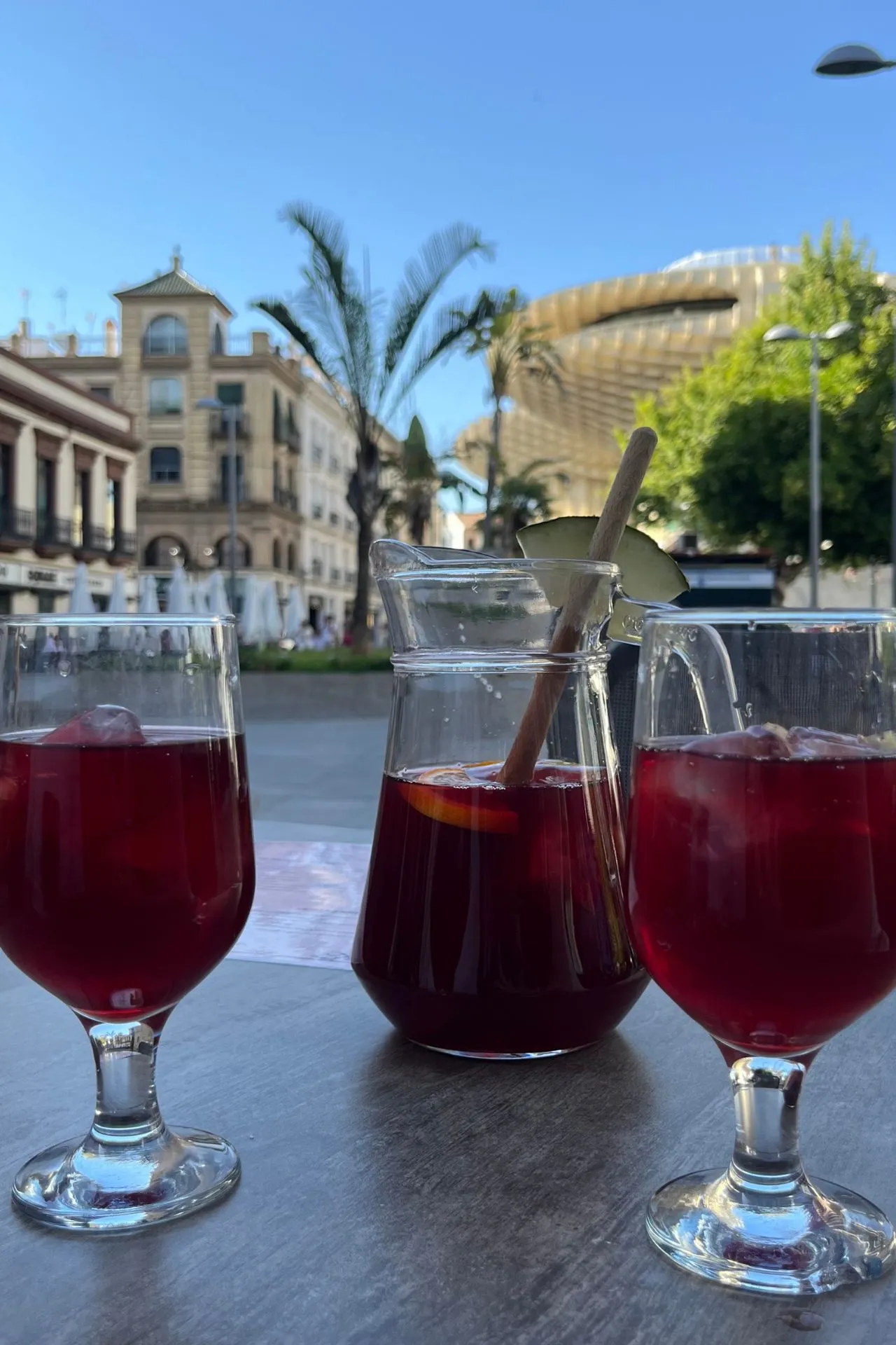 Twee glazen sangria en een karaf met schijfjes citrusvrucht op een terrastafel in Sevilla. Op de achtergrond zijn palmbomen, historische gebouwen en de houten structuur van de Metropol Parasol te zien.