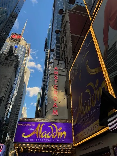 Het beroemde Broadway district in New York, met een groot "Amsterdam" bord en de reclame voor de musical Aladdin, verlicht door de iconische felle lichten van de stad.