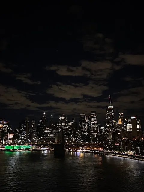 Avondfoto van de skyline van New York, met fonkelende lichtjes die weerspiegelen in het water, terwijl iconische wolkenkrabbers zoals het Empire State Building zich aftekenen tegen de donkere lucht.