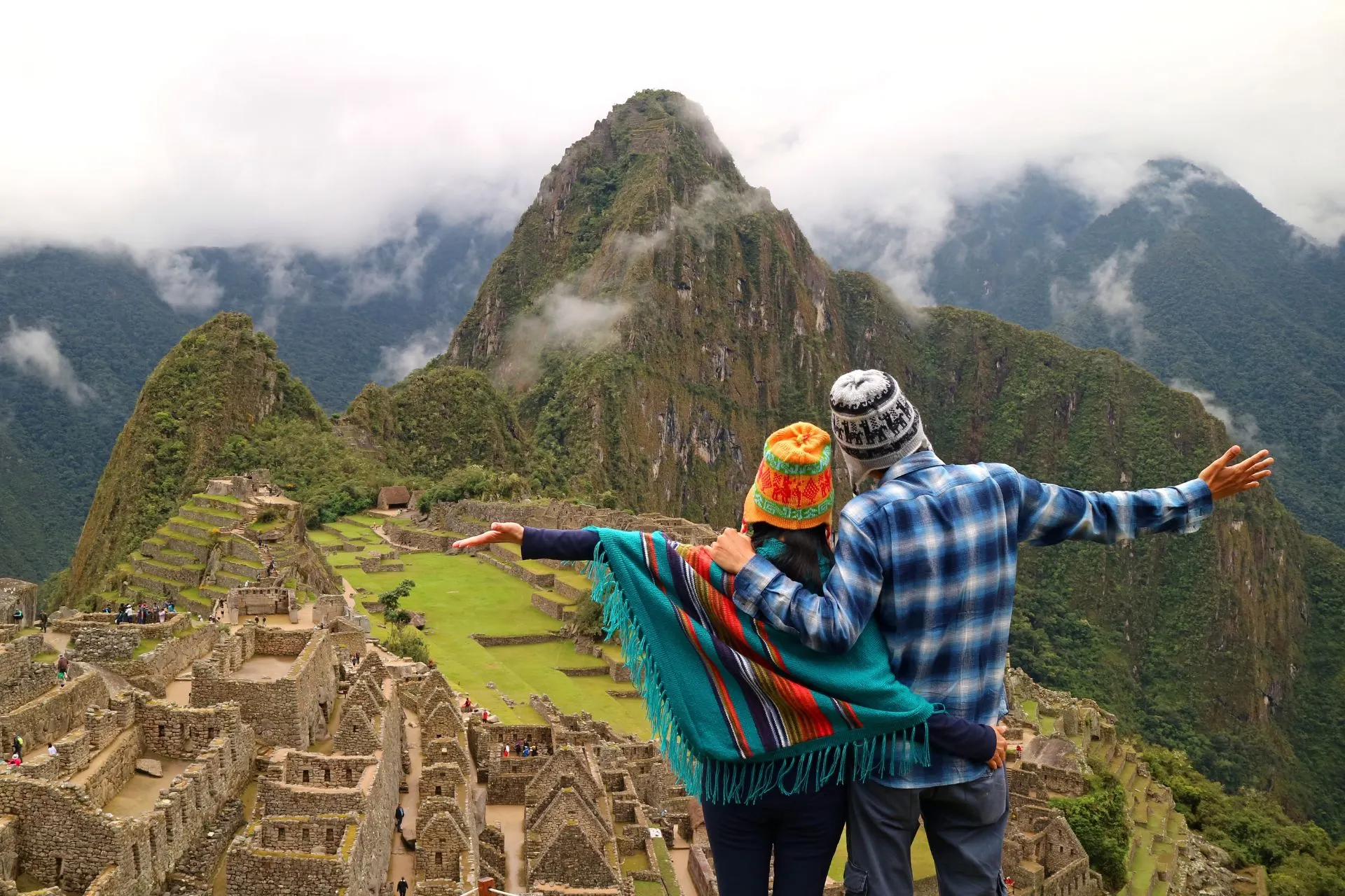 Een stel staat met open armen op een uitkijkpunt boven de oude Inca-stad Machu Picchu, omringd door groene bergen en zwevende wolken. Ze dragen kleurrijke Andes-mutsen en een poncho terwijl ze uitkijken over de ruïnes.