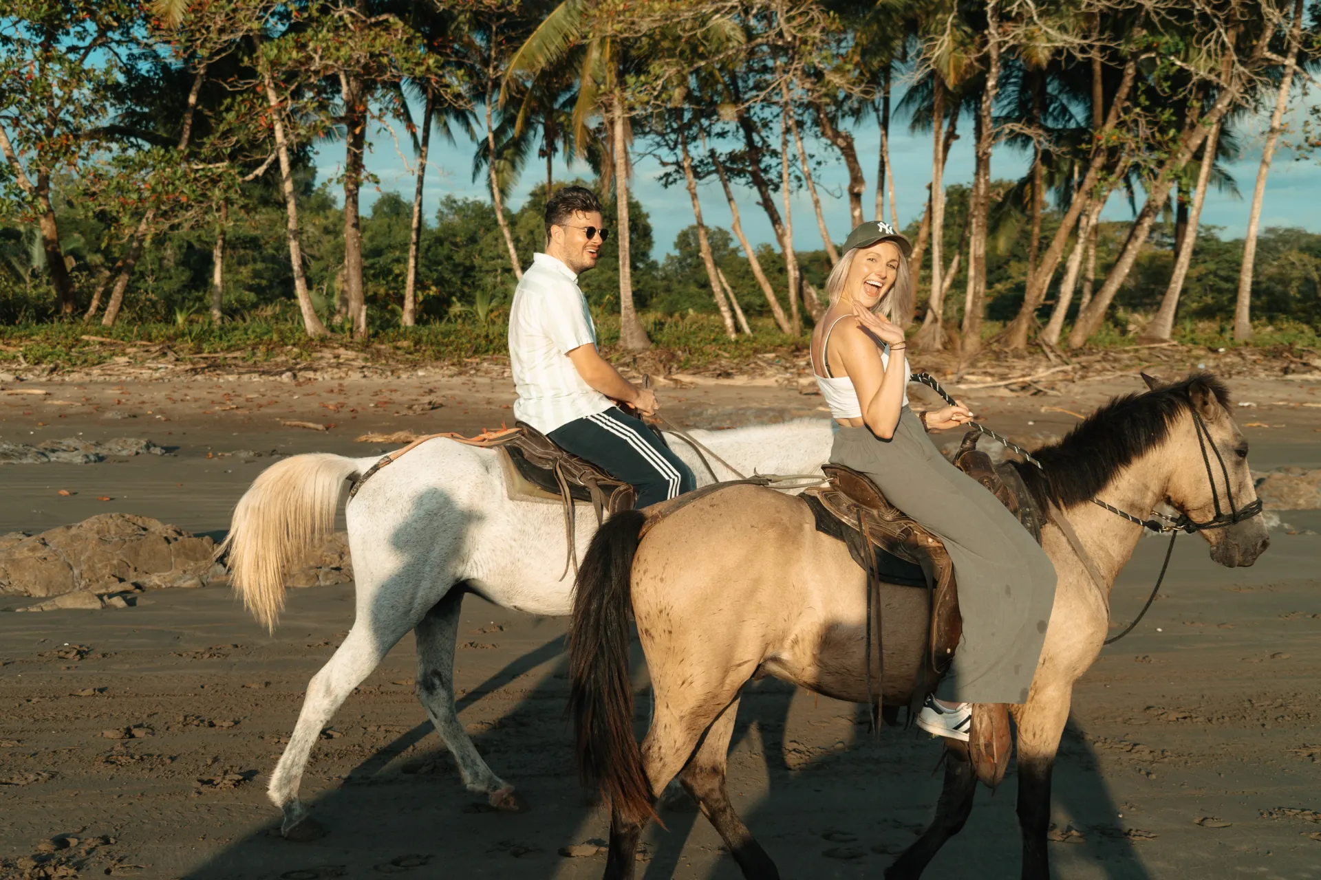 Twee mensen te paard op een strand in Santa Teresa, Costa Rica, lachend en genietend van een zonnige dag, met een rij palmbomen op de achtergrond.
