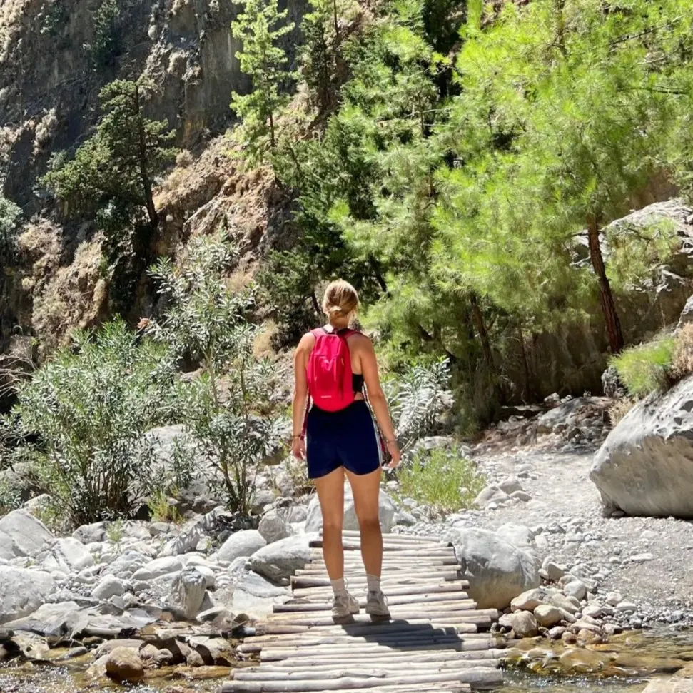 Vrouw met roze rugzak loopt over een houten brug over een beekje in de Samariakloof, omringd door groene bomen en ruige bergwanden.