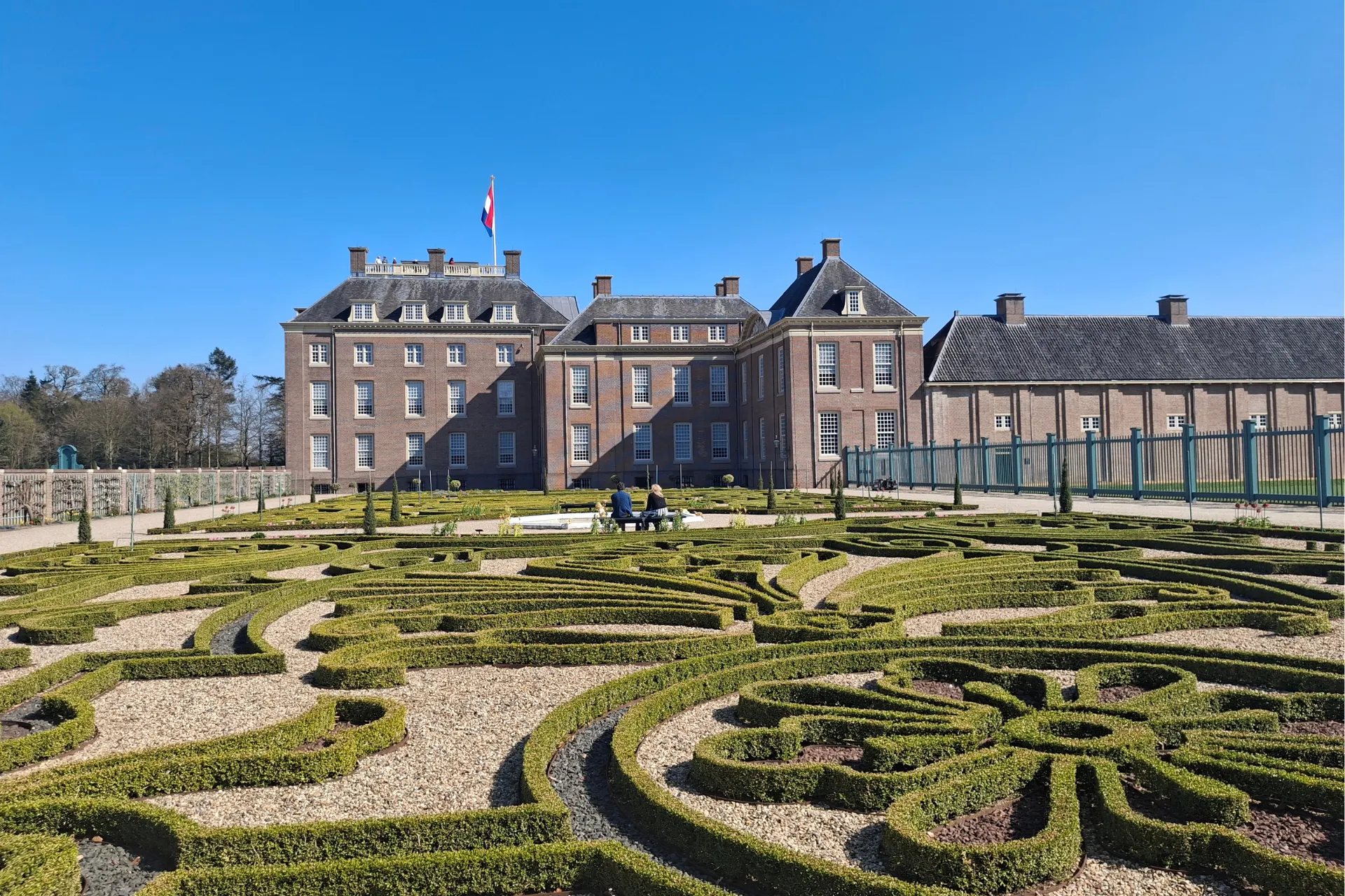 De tuin van Paleis Het Loo met sierlijke groen gevormde struiken en het paleis op de achtergrond.
